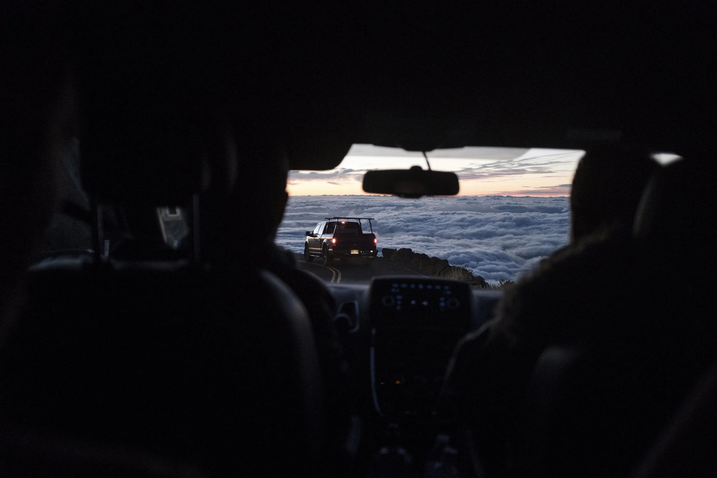 Vue depuis l'intérieur d'une voiture, montrant deux passagers regardant une voiture en marche sur une route de montagne au coucher du soleil, avec un ciel rempli de nuages.