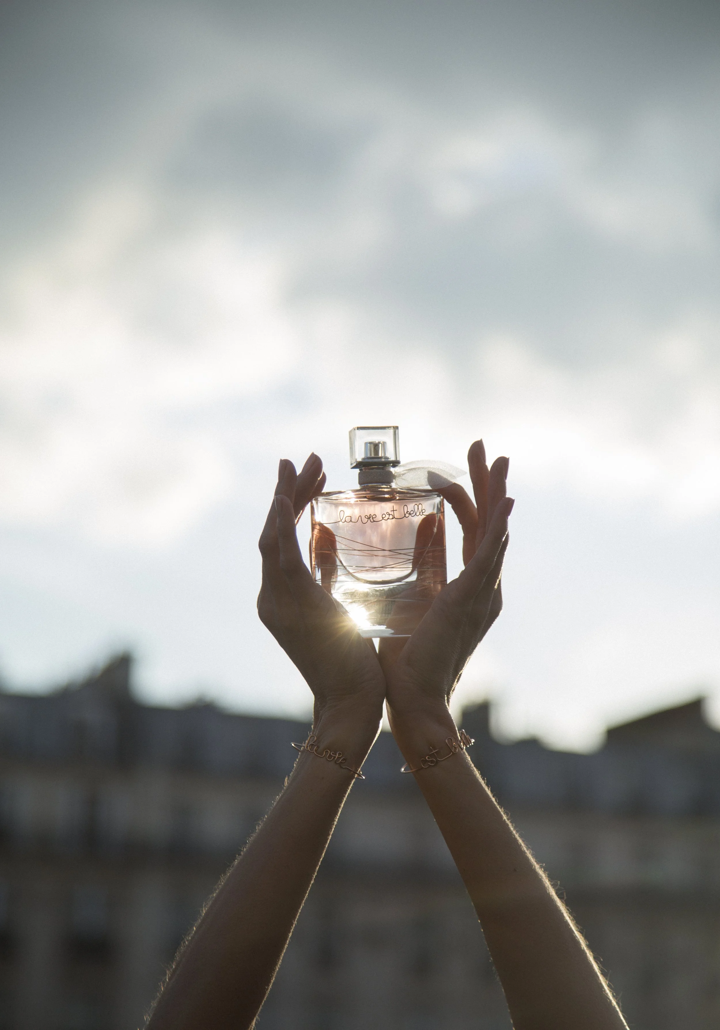 Une personne tient un flacon de parfum transparent contre le ciel nuageux, le soleil brille à travers le flacon.