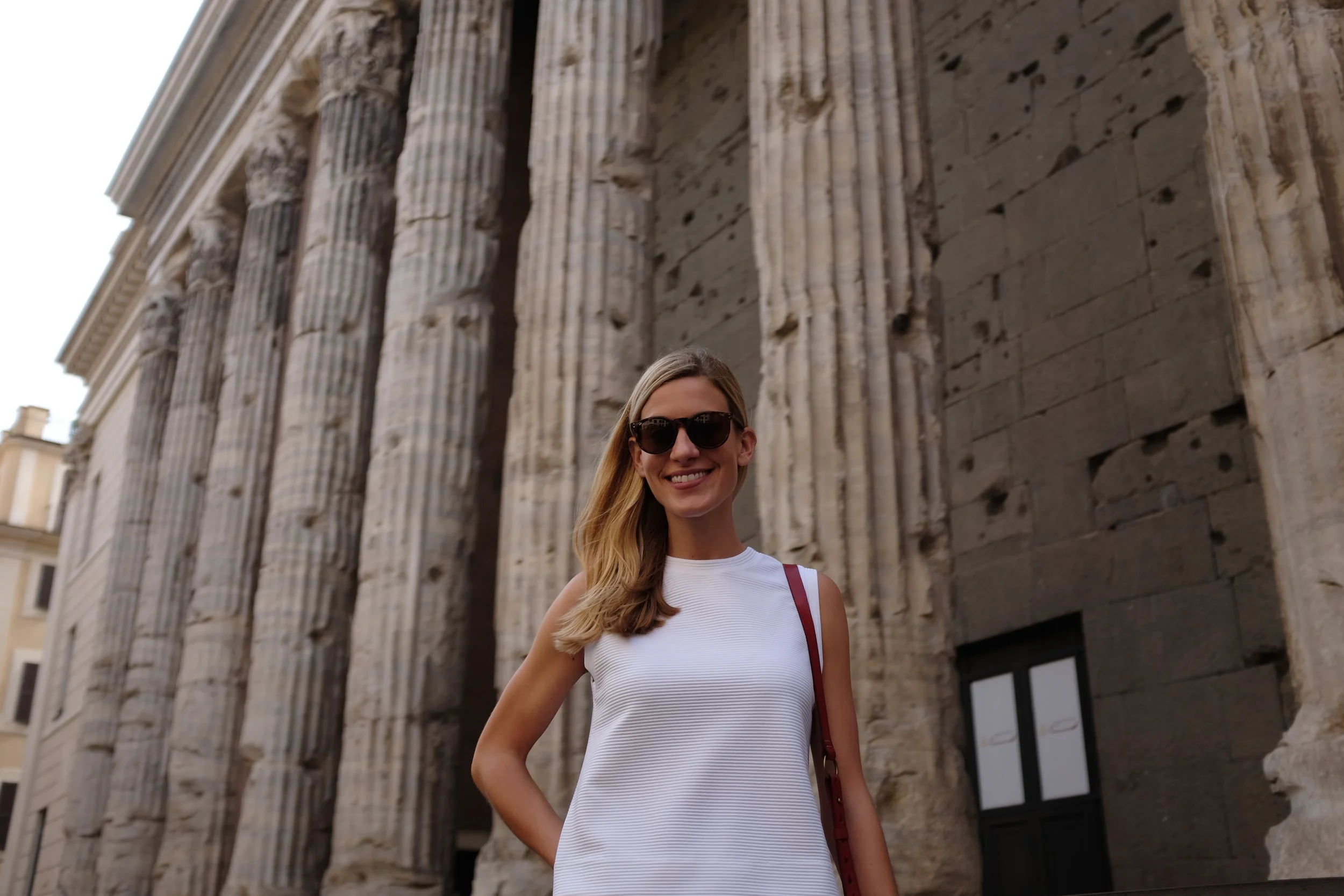 Une femme souriante portant des lunettes de soleil, une robe blanche et un sac rouge, devant des colonnes anciennes en pierre à Athènes.
