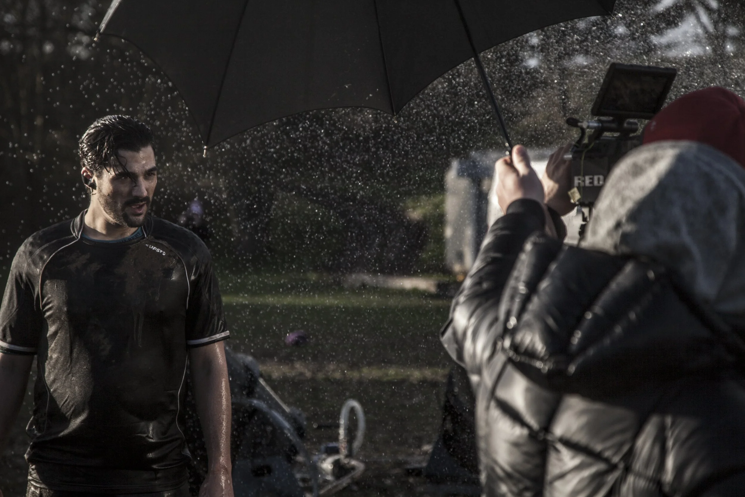 Une personne se faisant filmer par une équipe de tournage sous une pluie battante, tenant un parapluie noir.