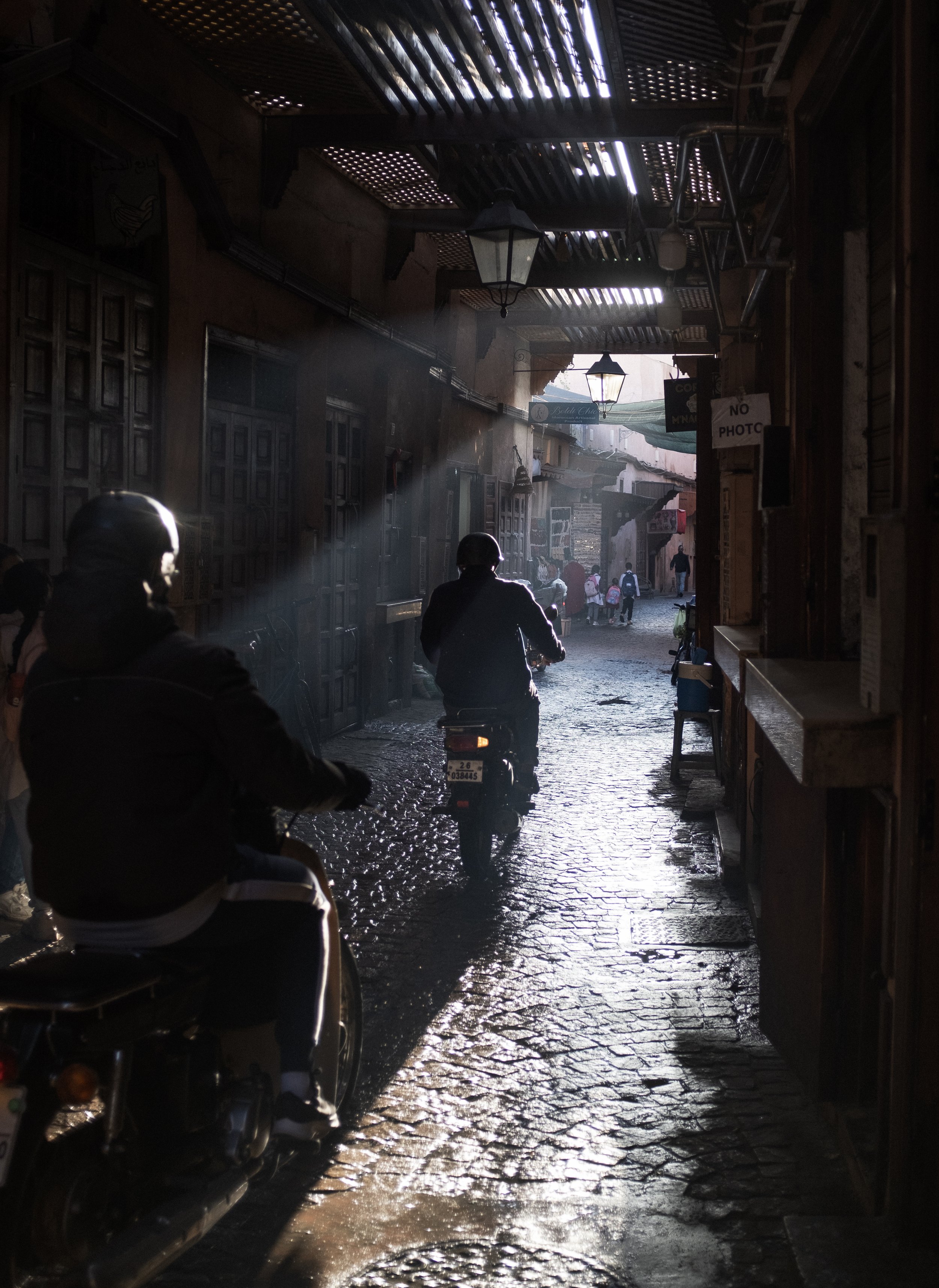 Vue d'une rue pavée étroite avec des motos et des personnes, où la lumière du soleil filtre à travers un toit en bois.