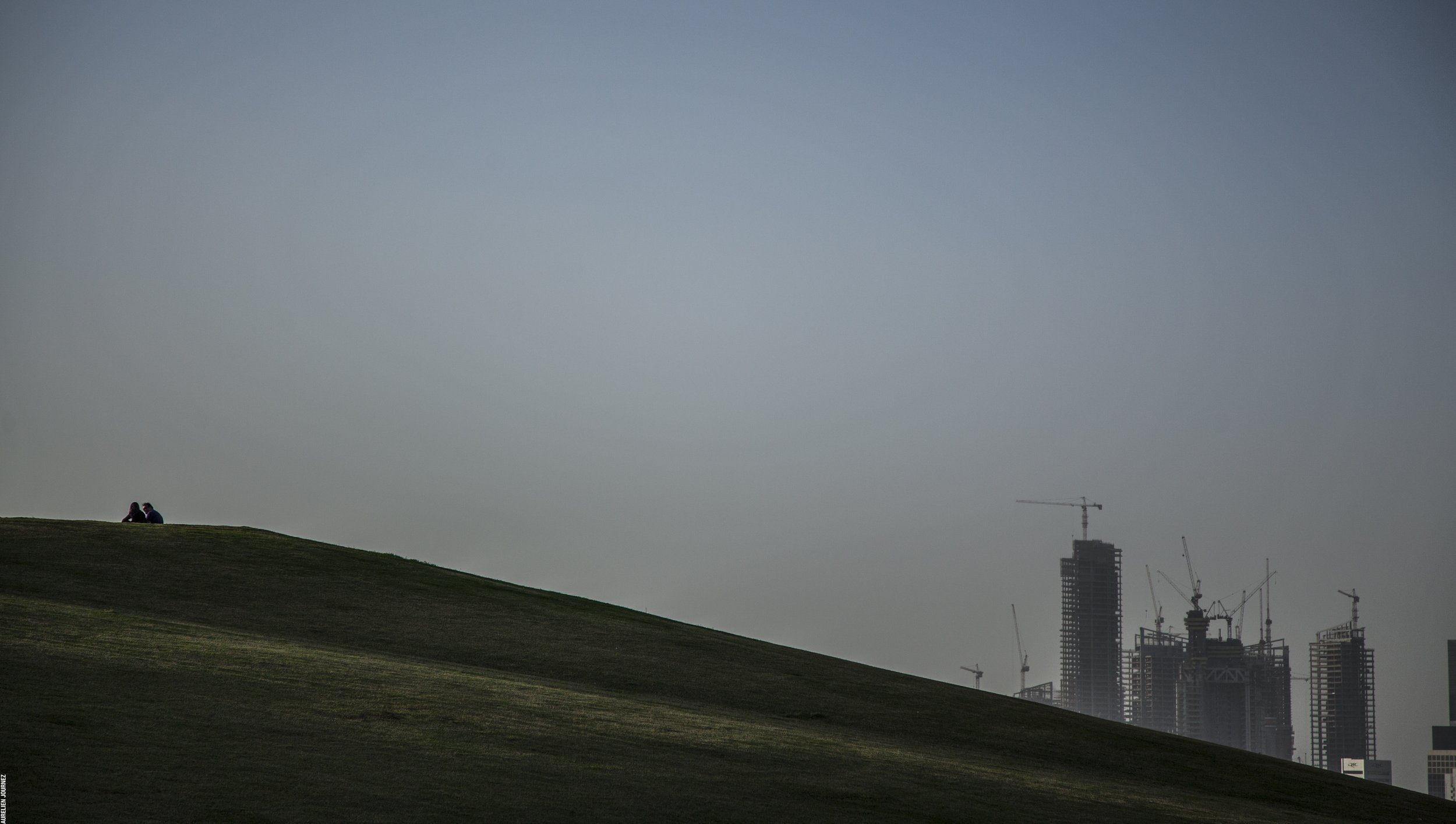 Deux personnes assises sur une colline verde qui regarde vers une ville en construction avec plusieurs grattes-ciel et grues, sous un ciel clair ou légèrement nuageux.