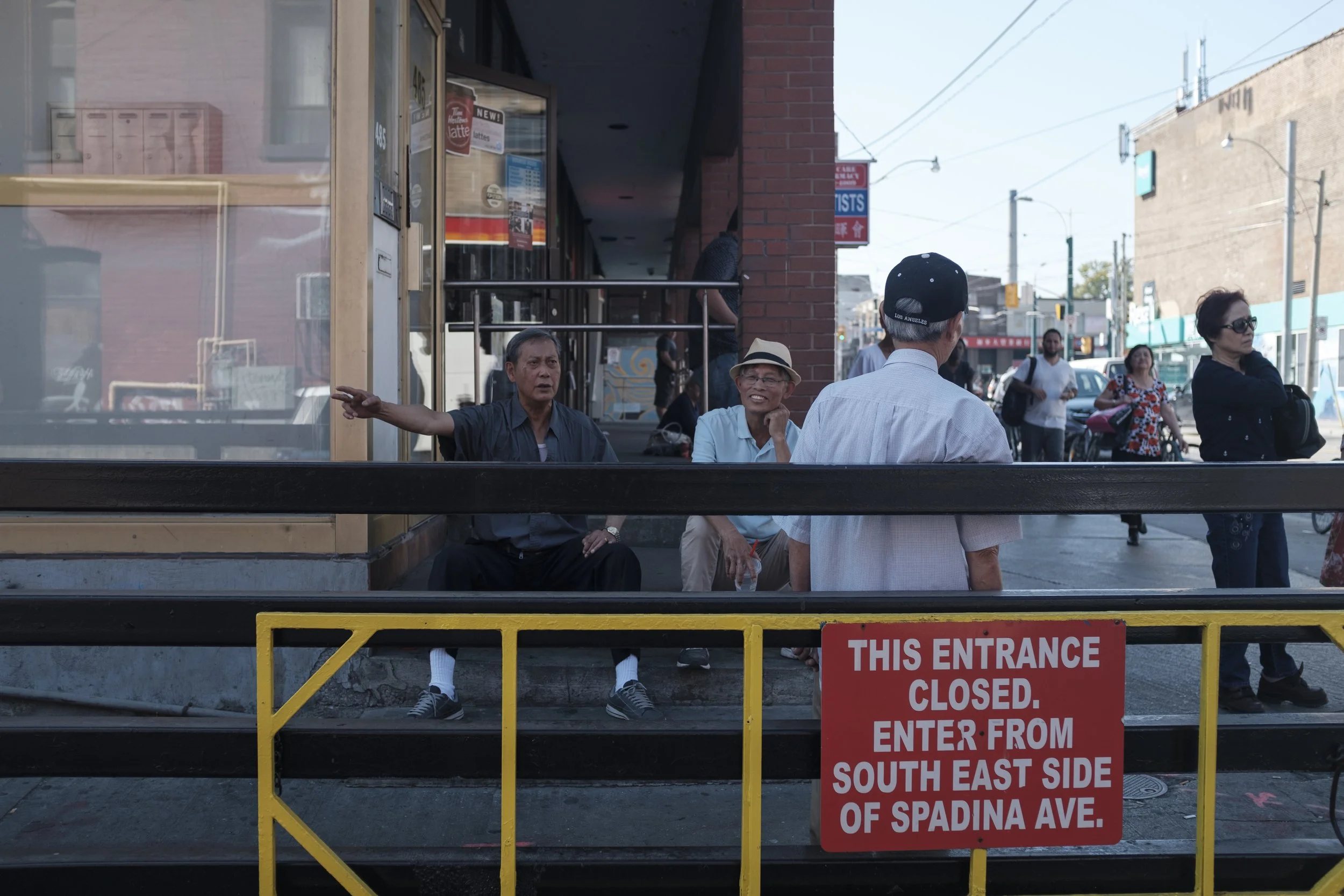 Groupe de personnes debout et assises à l'extérieur d'un bâtiment, avec un panneau rouge indiquant que l'entrée est fermée et qu'il faut entrer par le sud-est de Spadina Ave.