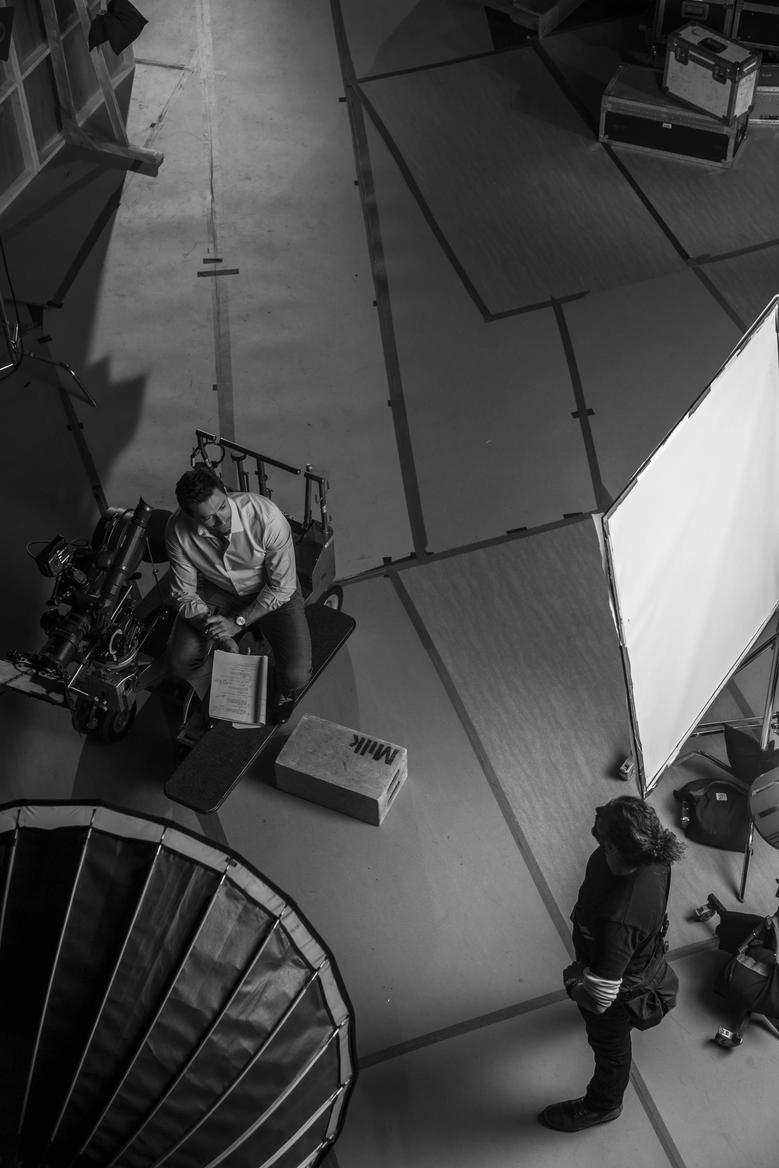 Prise de vue en top view d'un studio ou d'un théâtre avec deux personnes : une assise à côté d'une caméra et l'autre debout, entourées d’équipements de tournage et d’un grand écran de studio.