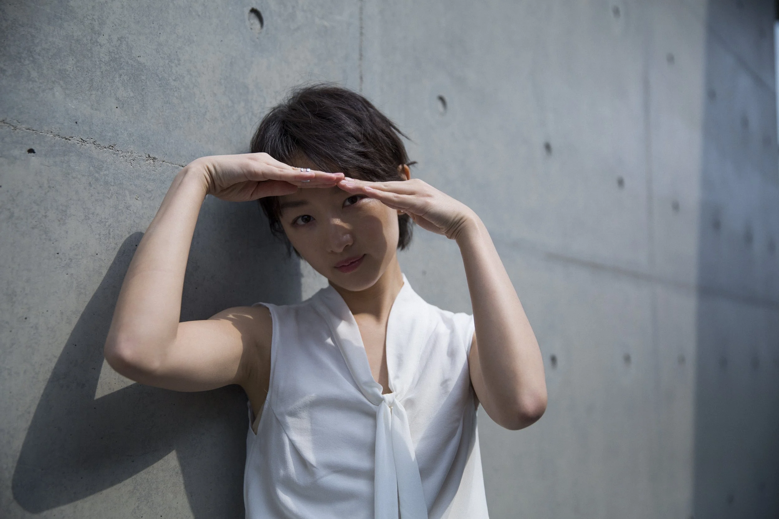 Une femme jeune avec une coiffure courte, portant une blouse blanche, se tenant contre un mur en béton et utilisant une main pour faire semblant de regarder au loin.