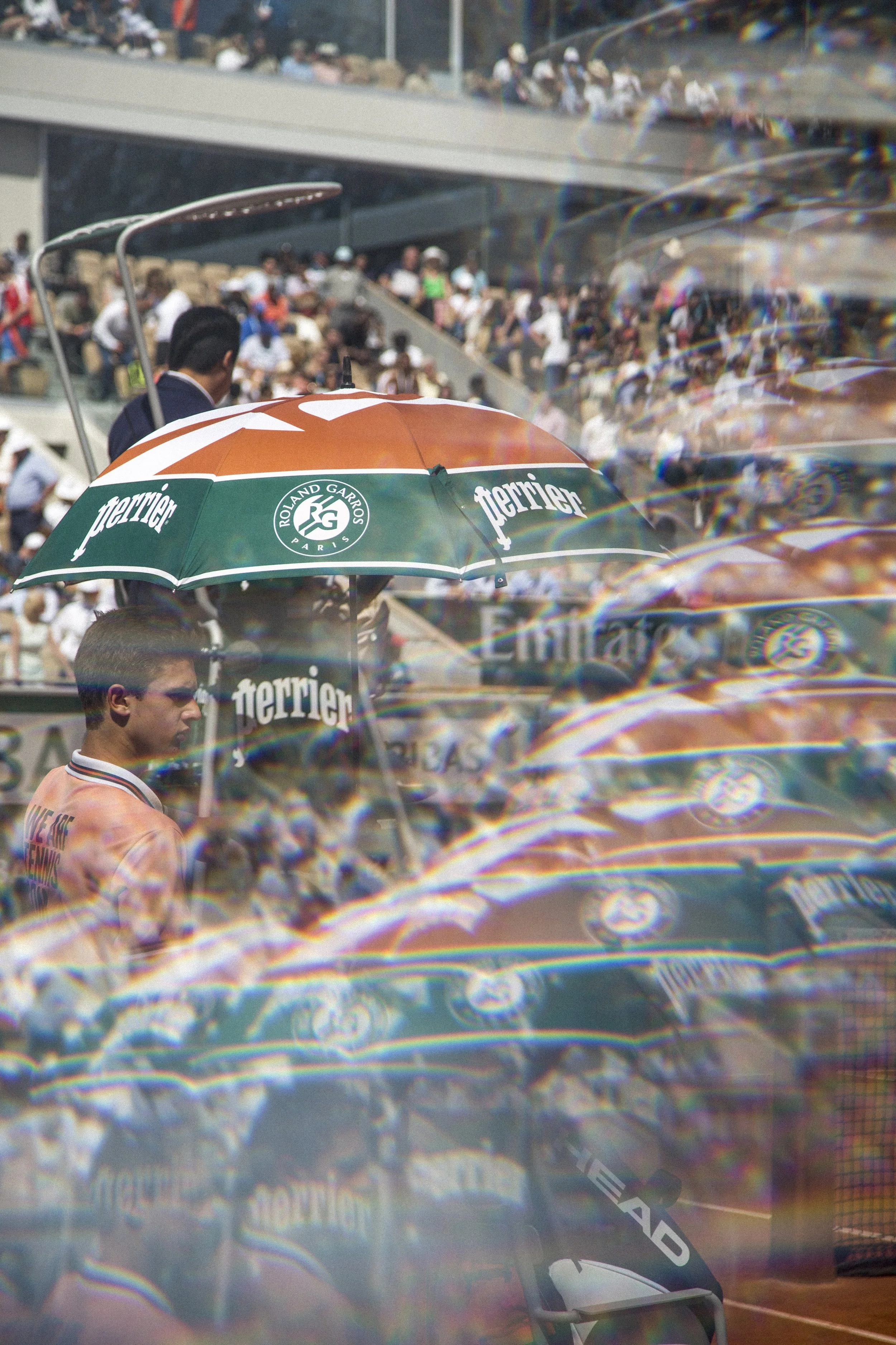 Une scène de tennis avec un spectateur sous un parapluie Ferrier, dans un stade rempli de spectateurs.