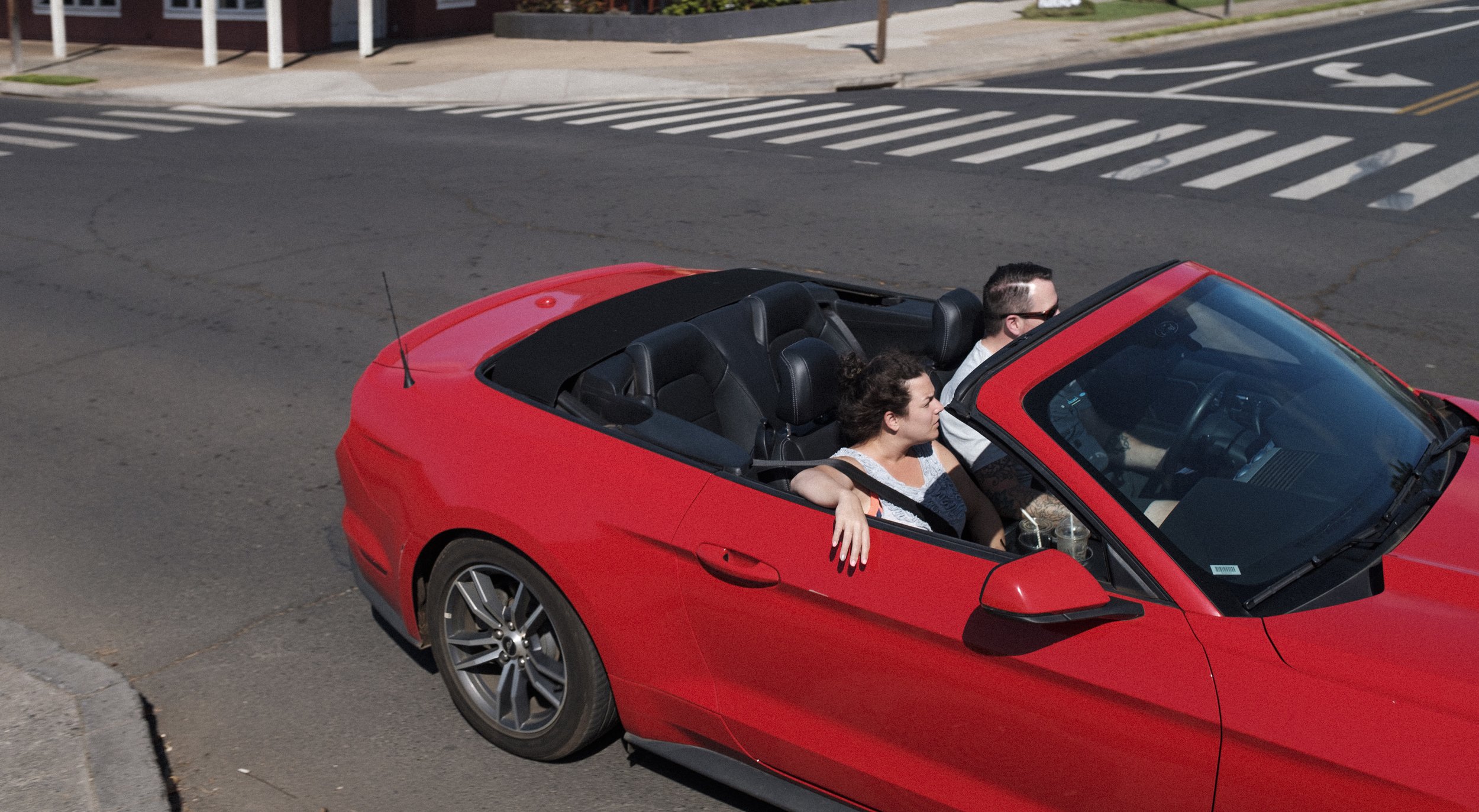 Une voiture décapotable rouge conduite par deux personnes, un homme et une femme, traversant une intersection urbaine en plein jour.