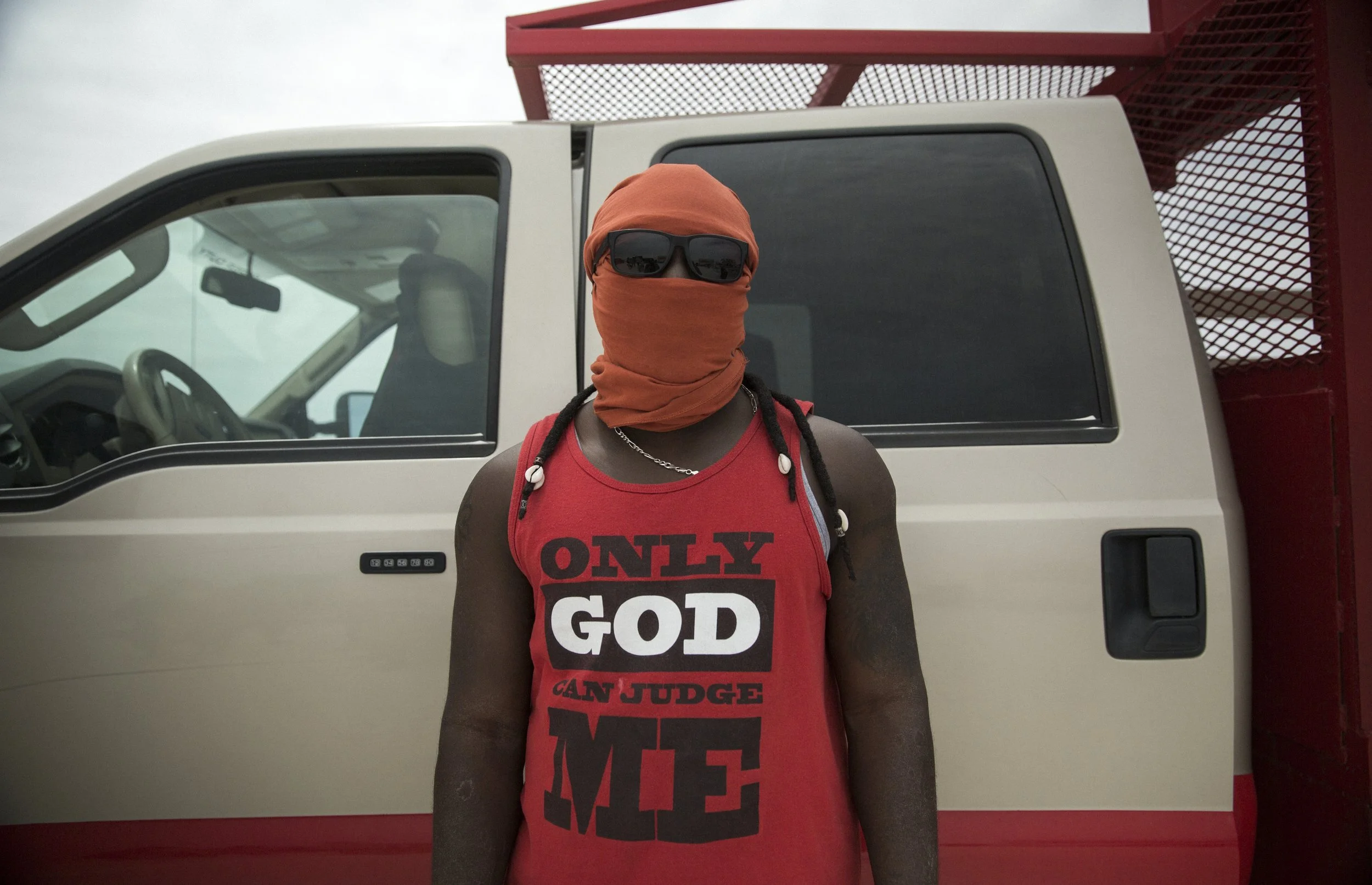 Un homme portant un bandeau en tissu orange, des lunettes de soleil, un t-shirt rouge avec le texte 'ONLY GOD CAN JUDGE ME', se tient devant un véhicule blanc. Le fond montre un ciel nuageux.
