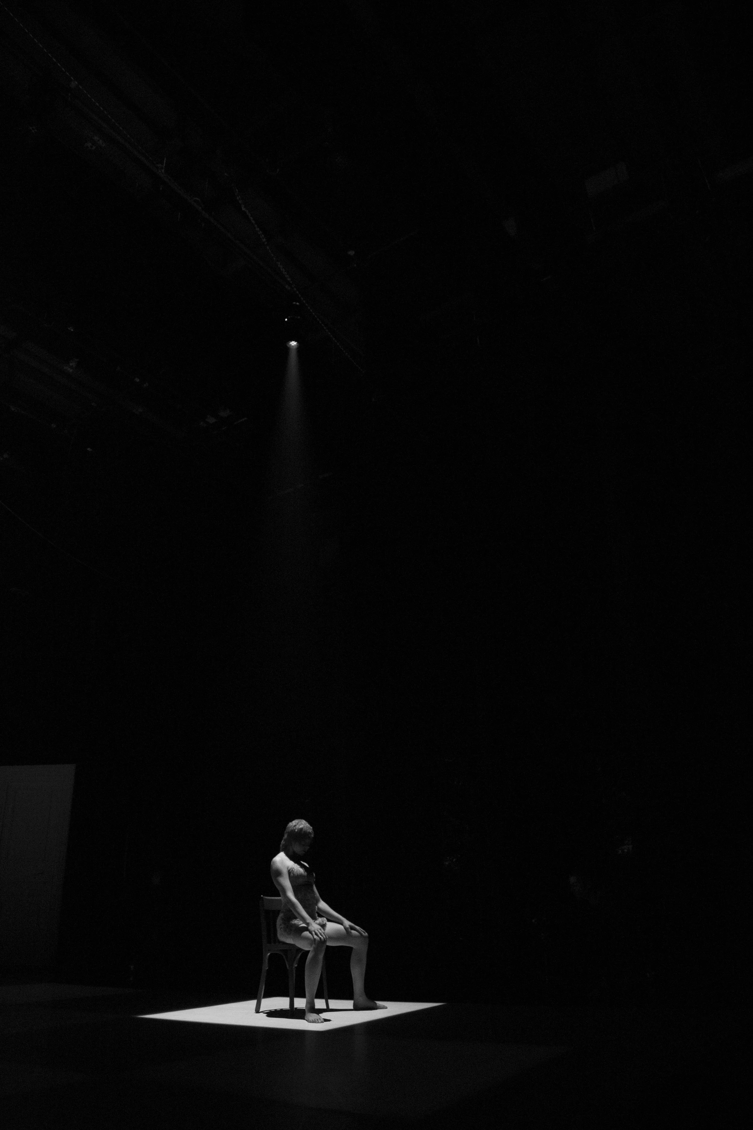 Une femme assise sur une chaise dans une pièce sombre, éclairée par un faisceau lumineux, créant un contraste élevé entre la lumière et l'obscurité.