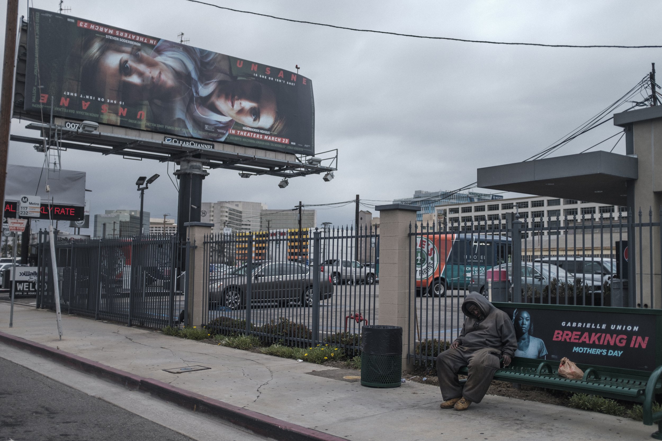 Un homme en vêtements amples assis sur un banc de parc, portant une capuche, dans une zone urbaine avec un parking, des affiches publicitaires et une clôture en fer. La scène est sombre avec un ciel nuageux.