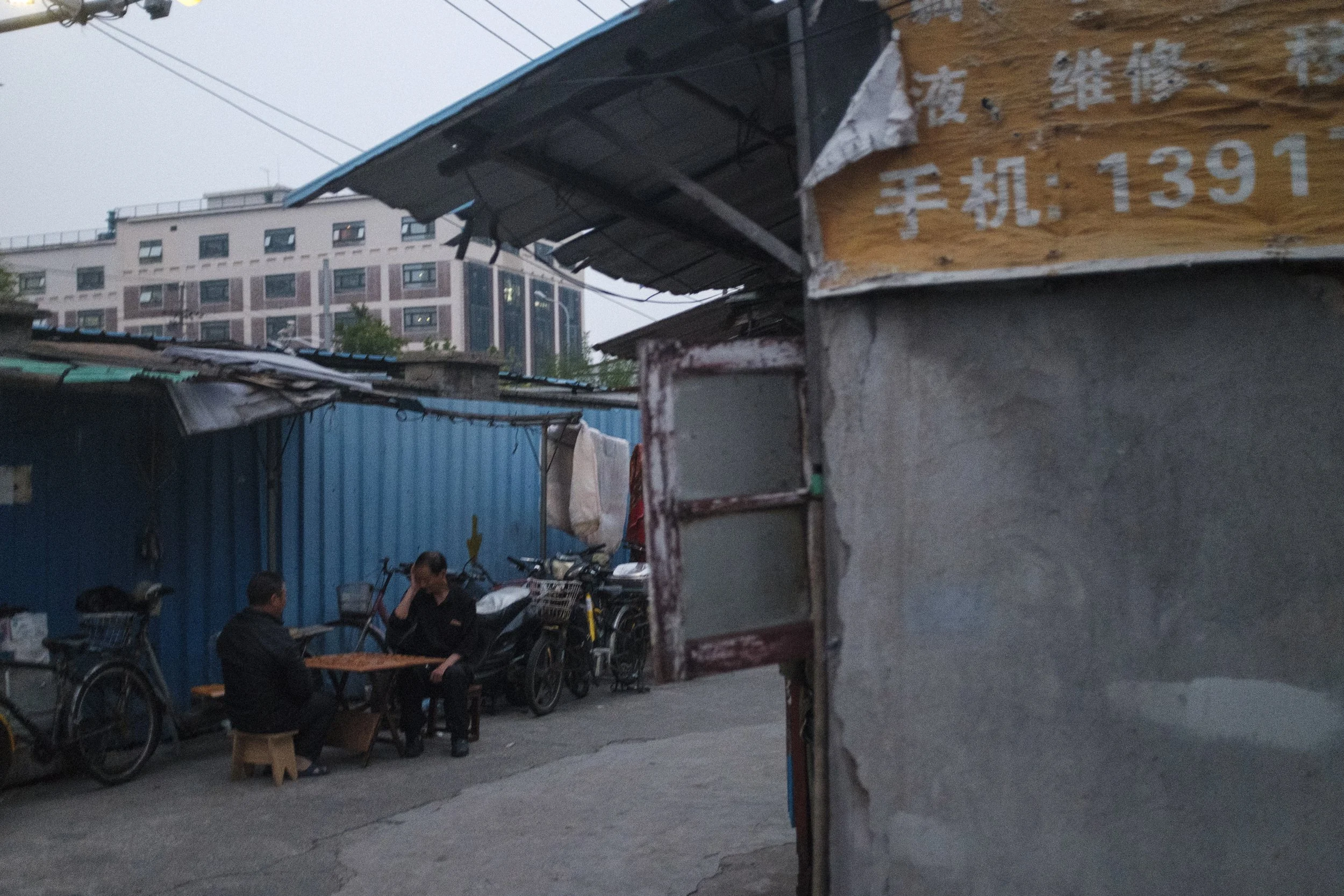 Deux personnes assises à une petite table dans une ruelle urbaine, entourées de vélos et de motos. En toile de fond, des bâtiments résidentiels et un mur en métal bleu. La lumière est faible, suggérant le début de soirée.