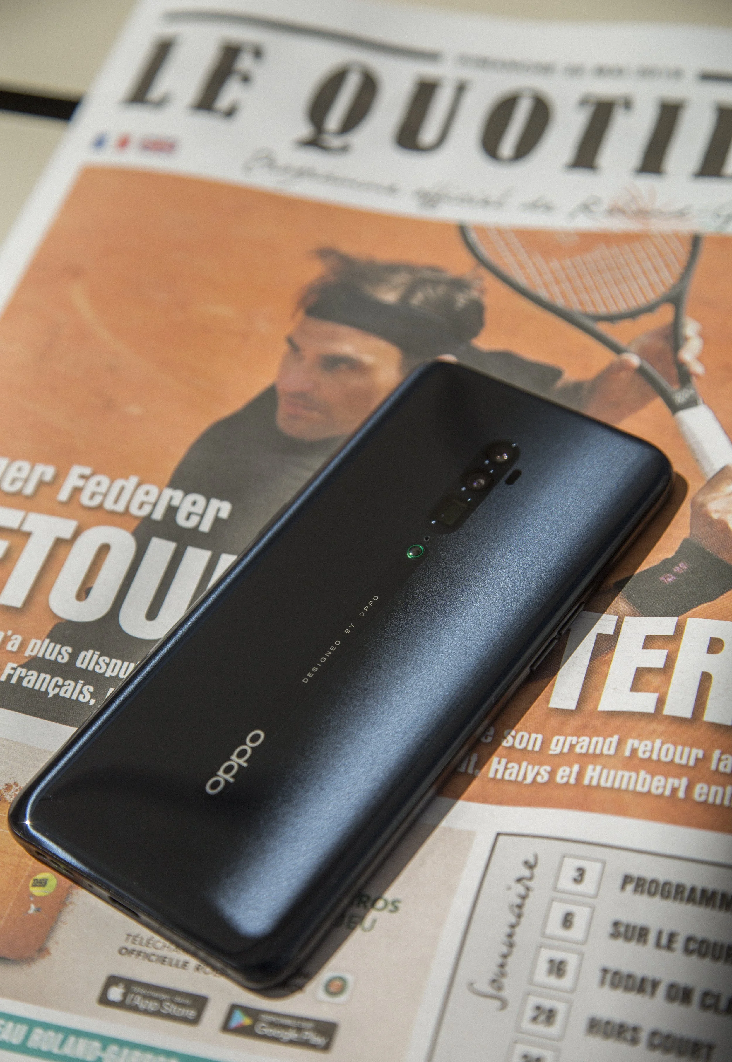 Un téléphone portable noir posé sur un journal. Le journal affiche une image d’un homme avec un bandeau à la tête jouant au tennis, avec du texte en français.