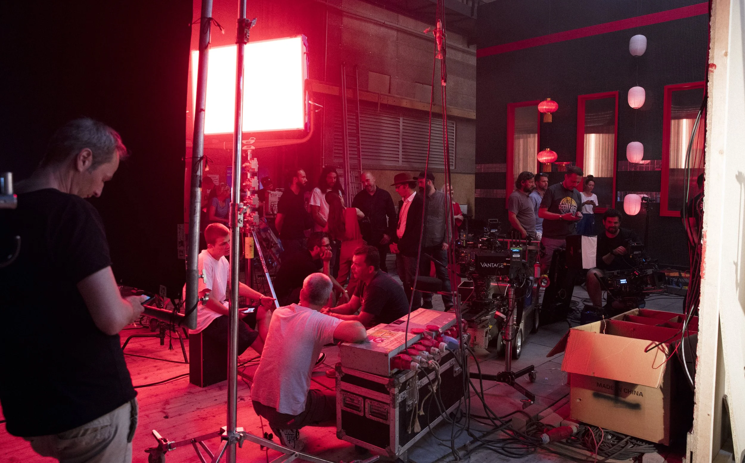 Coulisses d'un tournage ou d'une production vidéo avec plusieurs personnes autour d'équipements caméra, lumière, et câbles, dans un espace intérieur décoré avec des lanternes rouges et blanches.