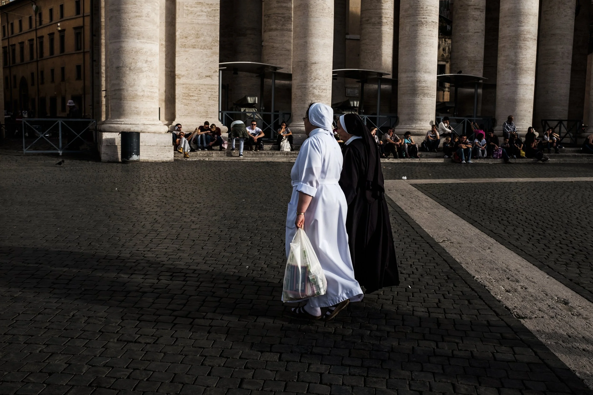 Deux religieuses, une en blanc et une en noir, marchent côte à côte sur une place pavée, avec des gens assis en arrière-plan devant une grande structure avec des colonnes.