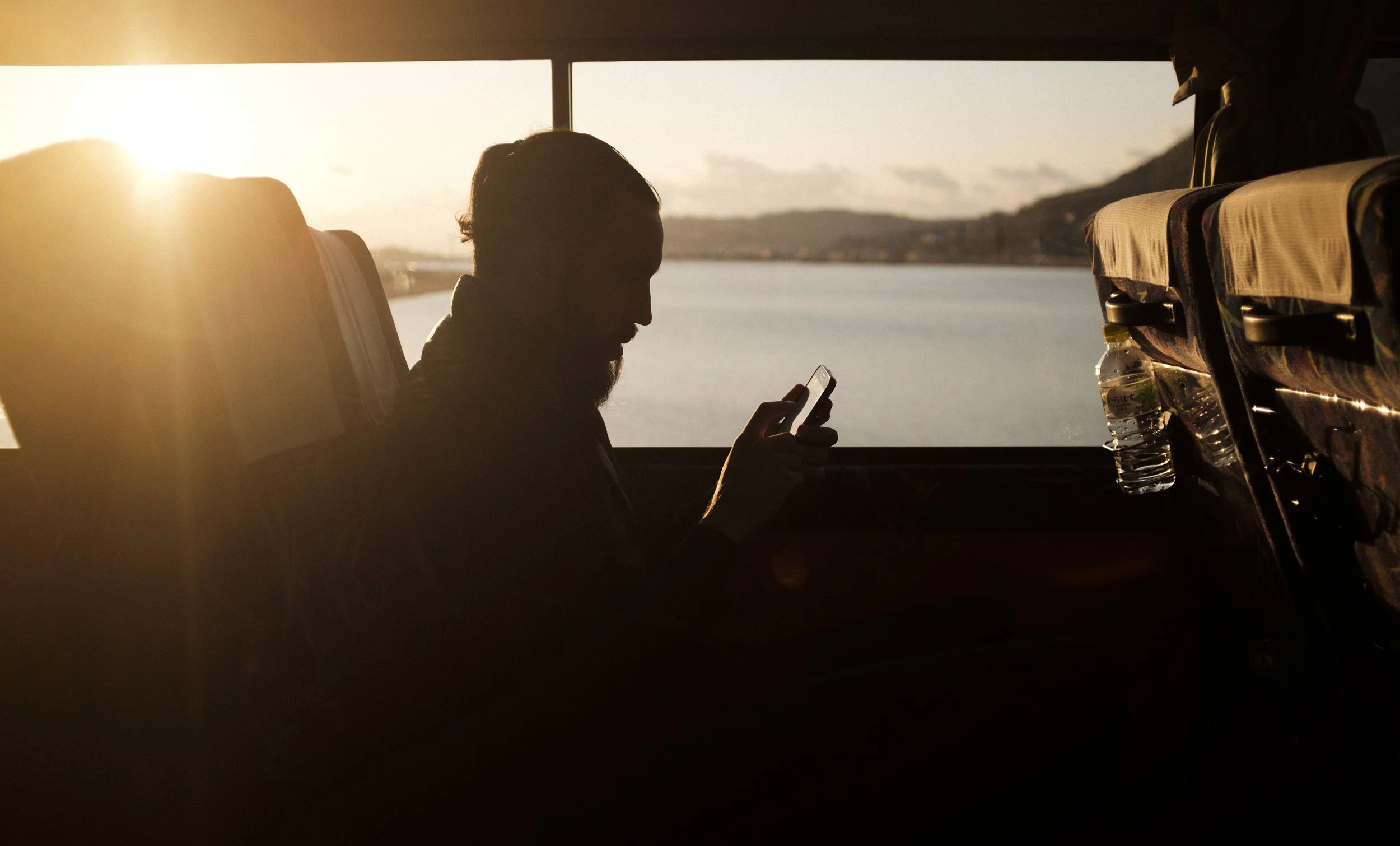 Une personne silhouette utilisant un téléphone dans un bus au coucher du soleil, avec un paysage de rivière et de montagnes en arrière-plan.