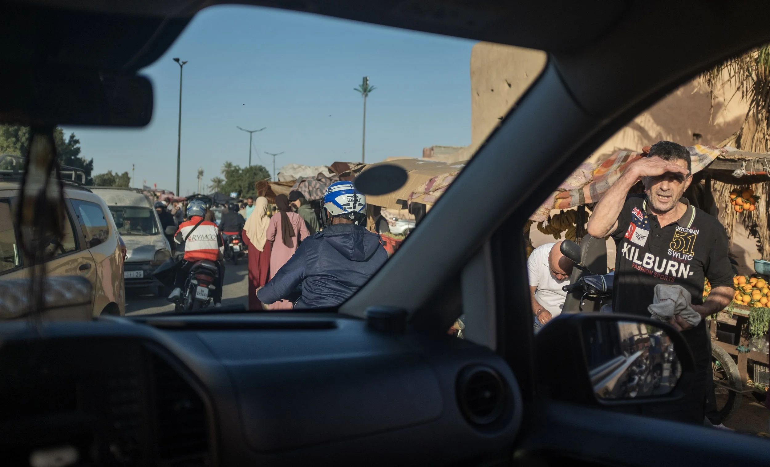 Un marché animé avec des gens et des véhicules, une personne en t-shirt noir avec des inscriptions, un homme portant un casque de moto, et des étals de fruits en arrière-plan.