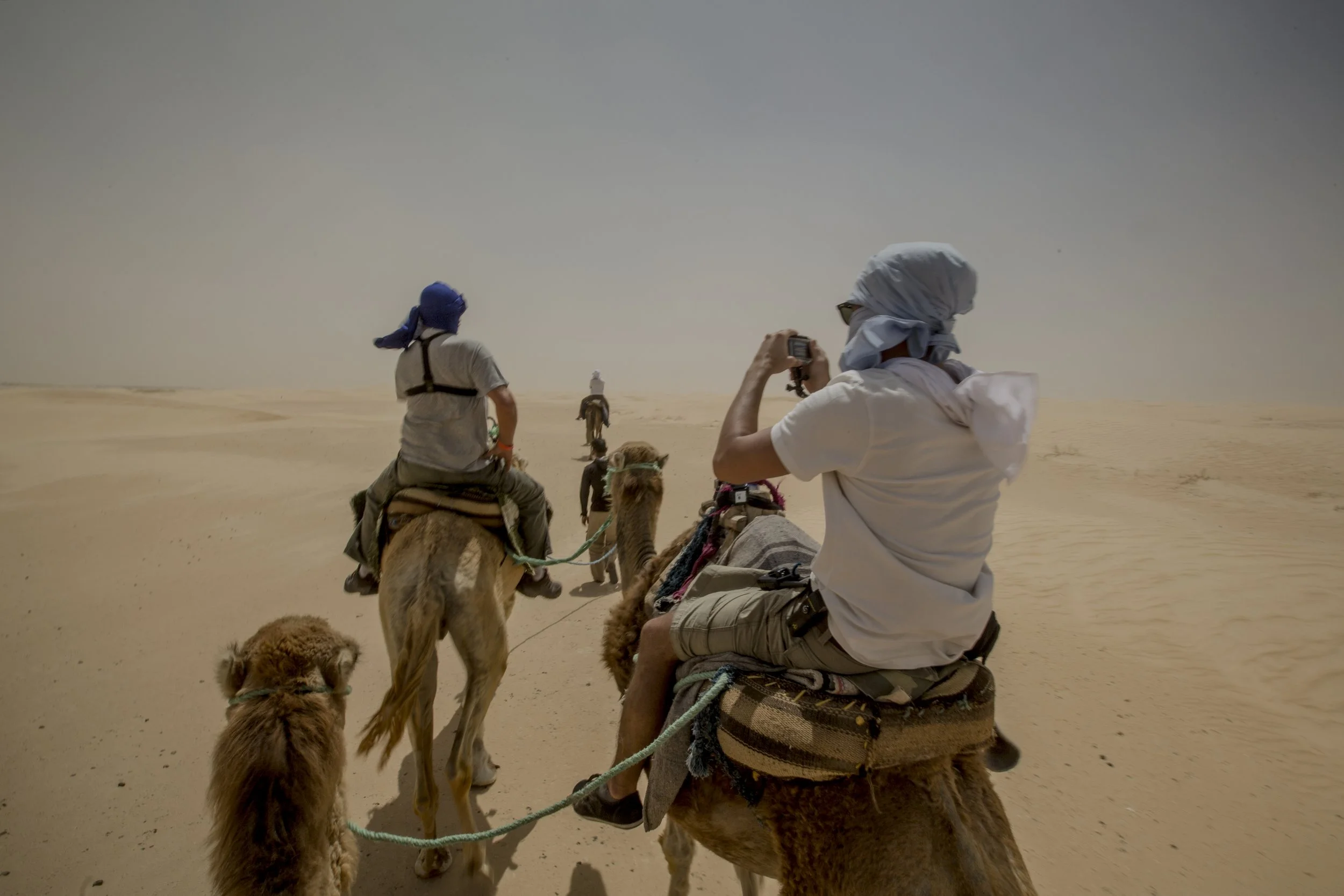 Groupe de touristes à dos de chameau dans un désert.
Un homme en blanc avec un foulard sur la tête prend une photo avec son téléphone, d'autres personnes sont visibles en arrière-plan, aussi à dos de chameau.