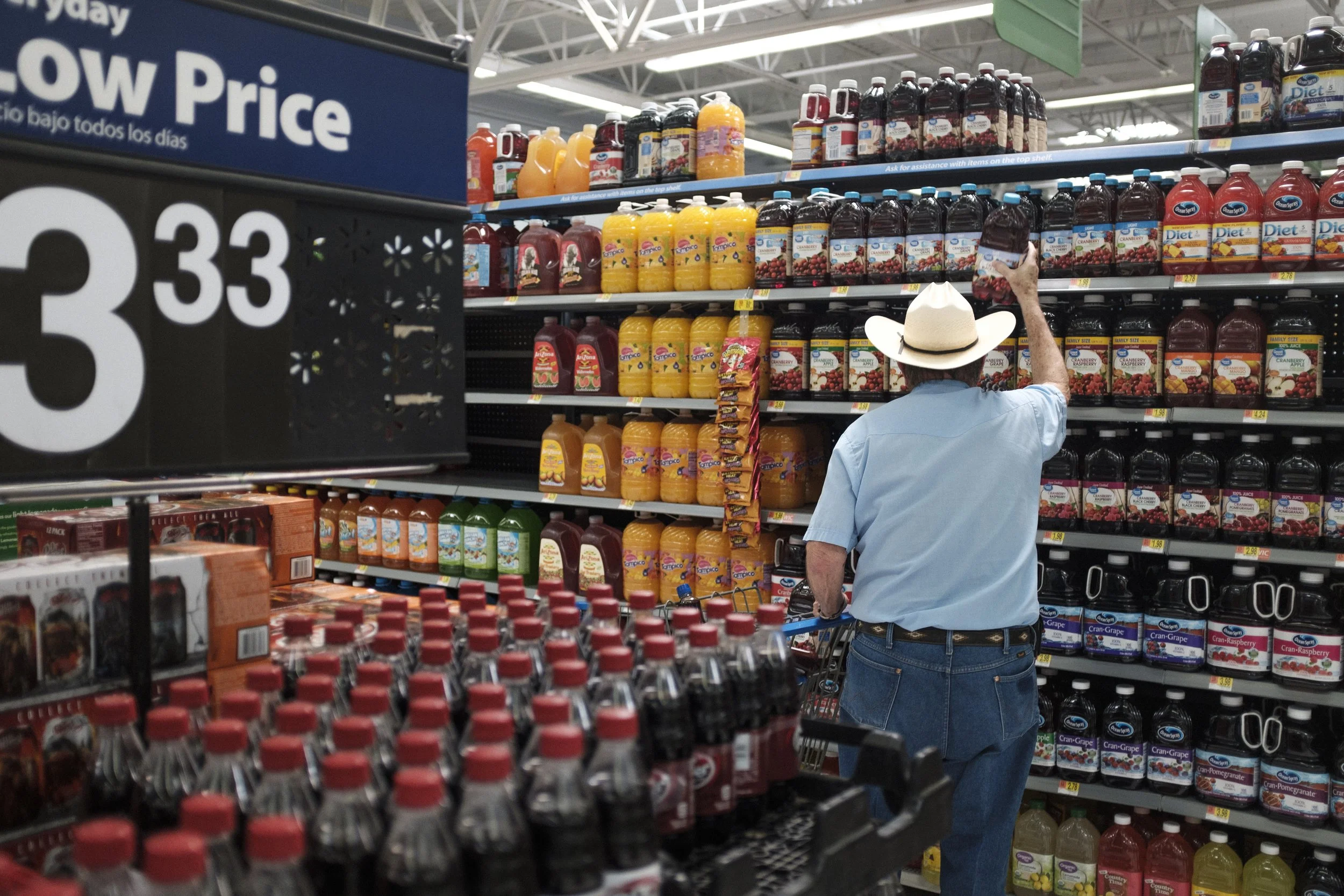 Un homme portant un chapeau blanc et une chemise bleue achète des jus de fruits en supermarché, face à des étagères remplies de bouteilles de jus de différentes saveurs.