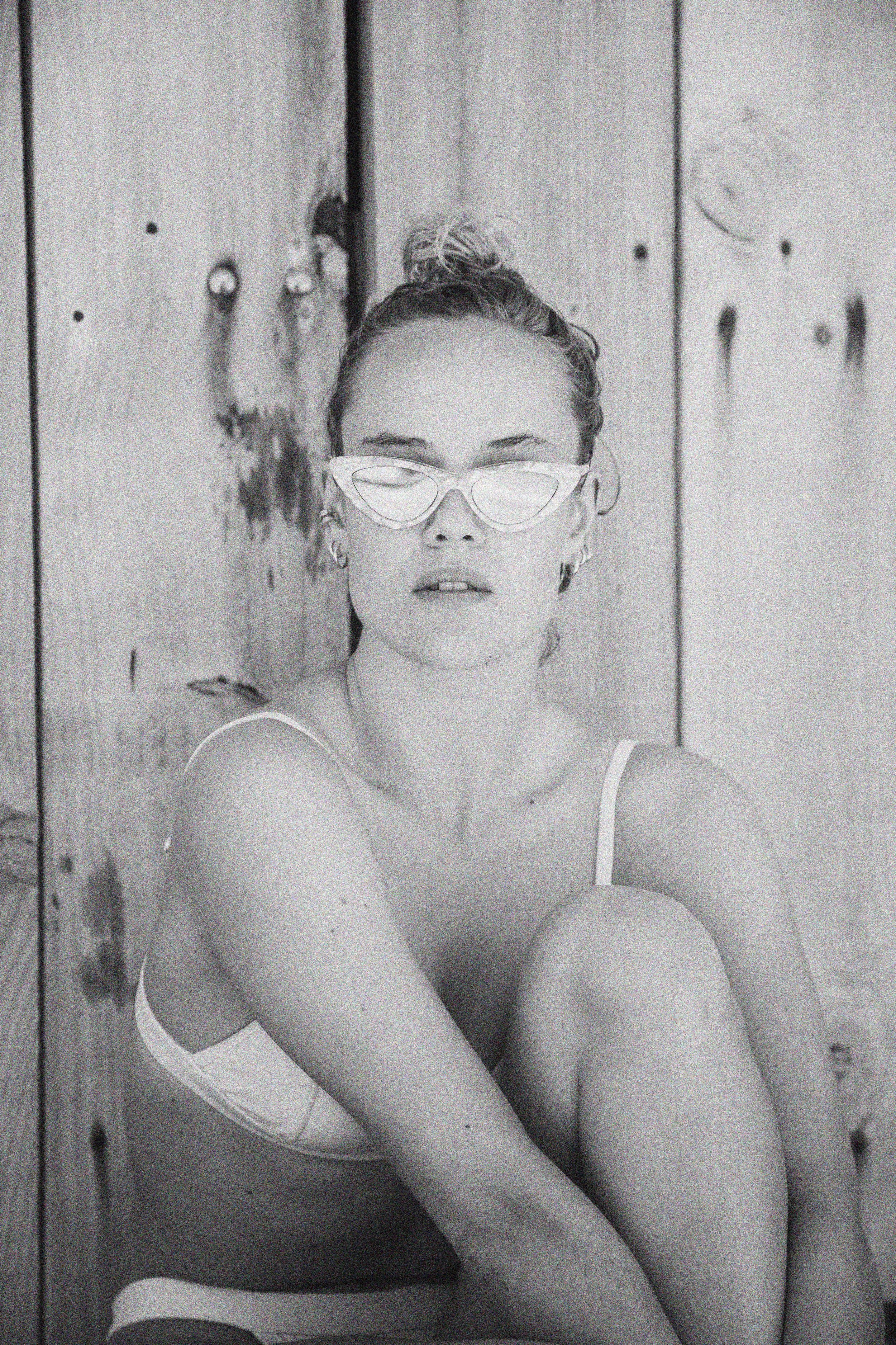 Portrait en noir et blanc d'une femme avec des lunettes de soleil, assise contre un mur en bois, regardant à l'objectif.