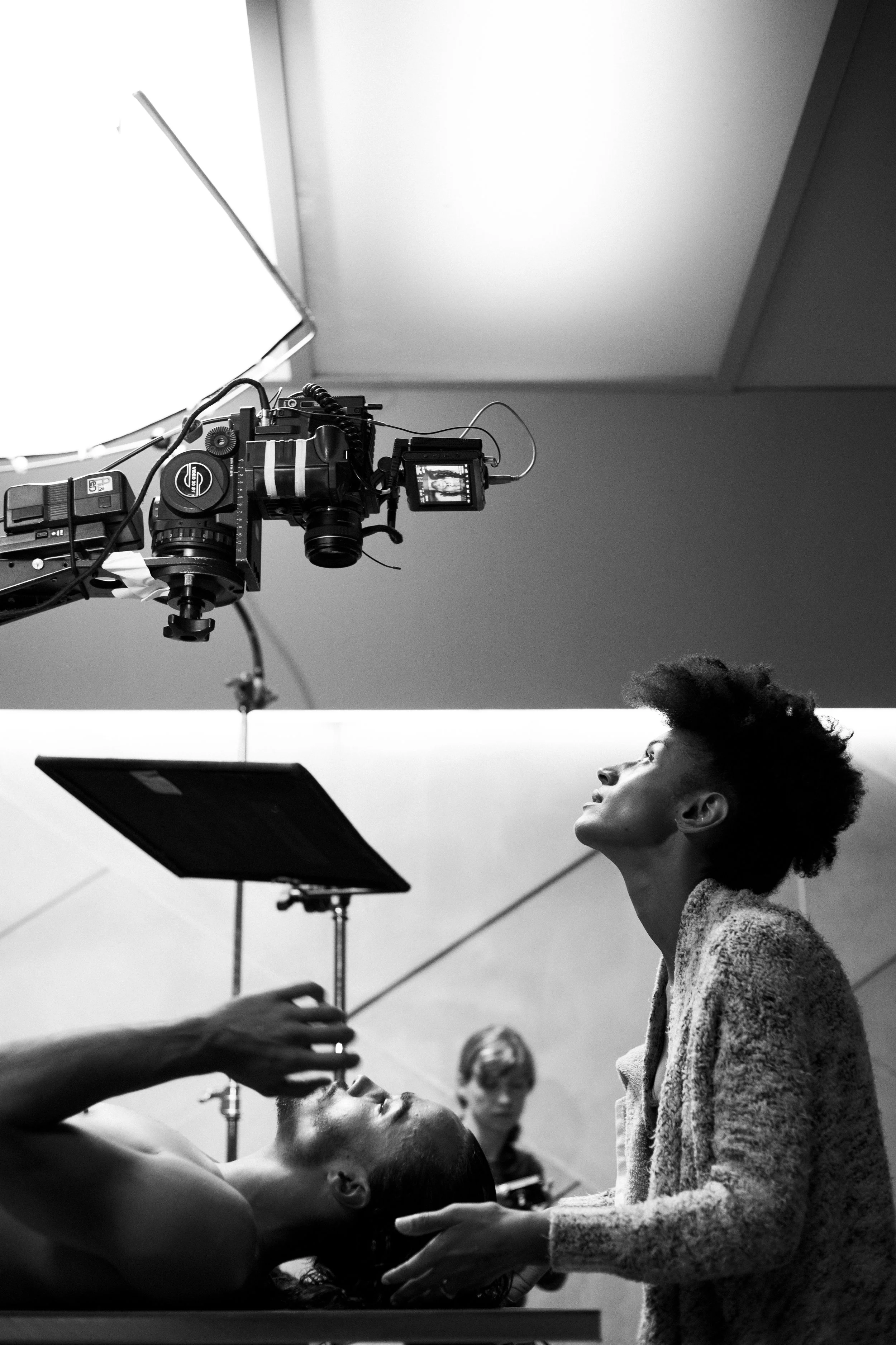 Une séance de tournage en noir et blanc, avec une femme aux cheveux bouclés assise et une autre personne en train de la maquiller ou de la préparer, sous un appareil photo professionnel suspendu au plafond, avec une personne en arrière-plan en train 