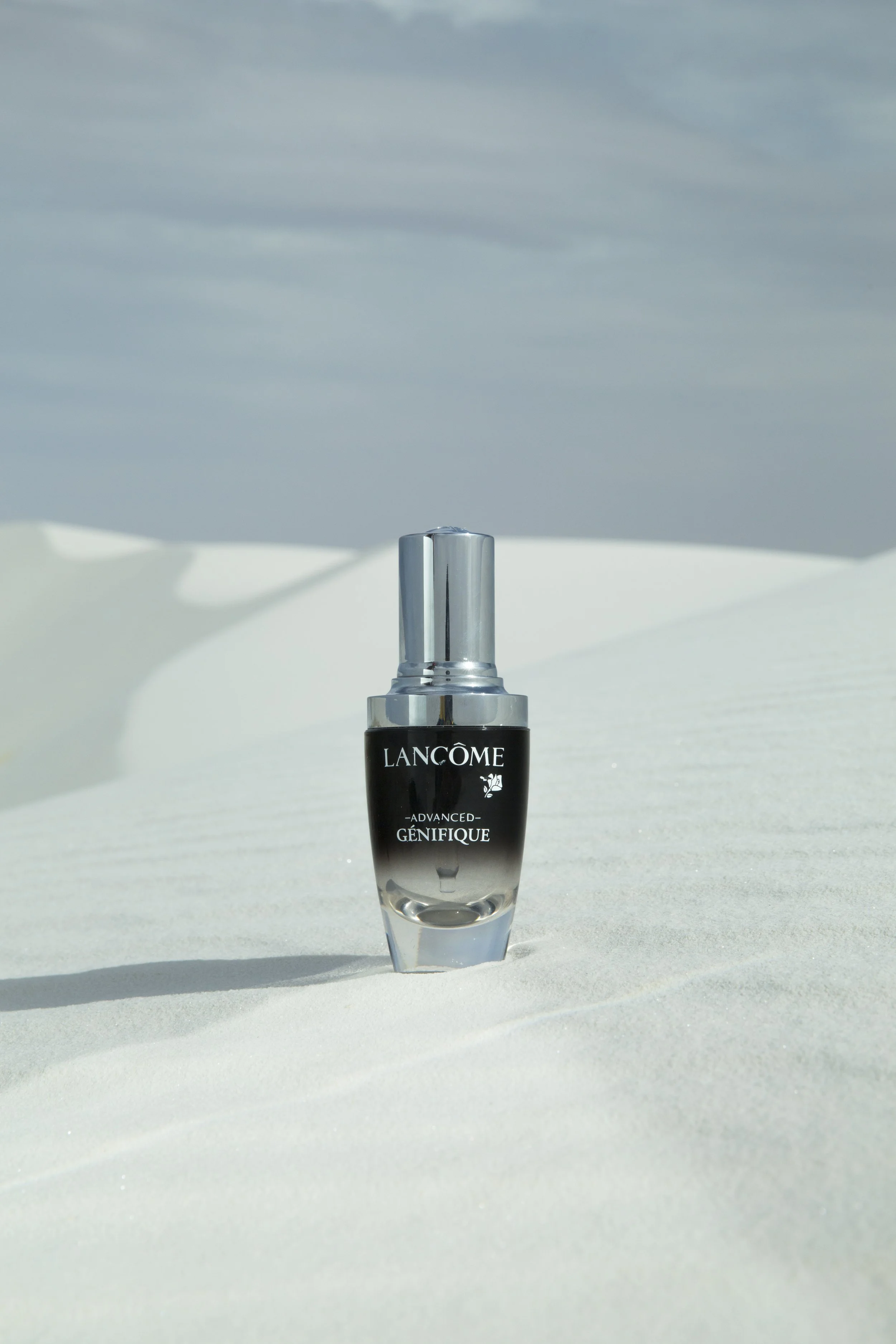 Flacon de parfum Lancôme Advanced Génifique posé sur du sable blanc dans un paysage aride avec un ciel nuageux.