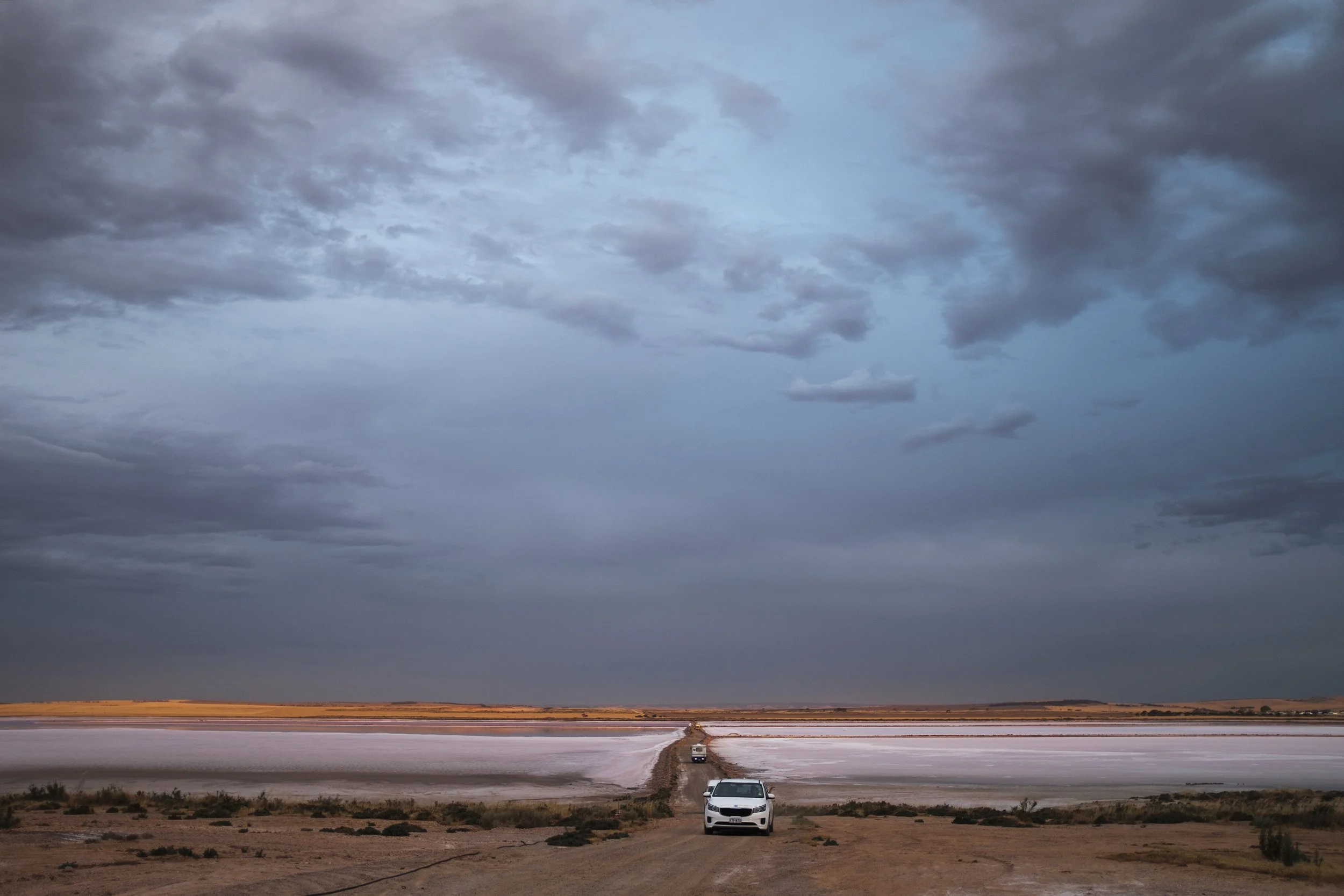 Voiture blanche sur une route de terre dans un paysage désertique avec un lac salé et des montagnes à l'horizon sous un ciel nuageux.