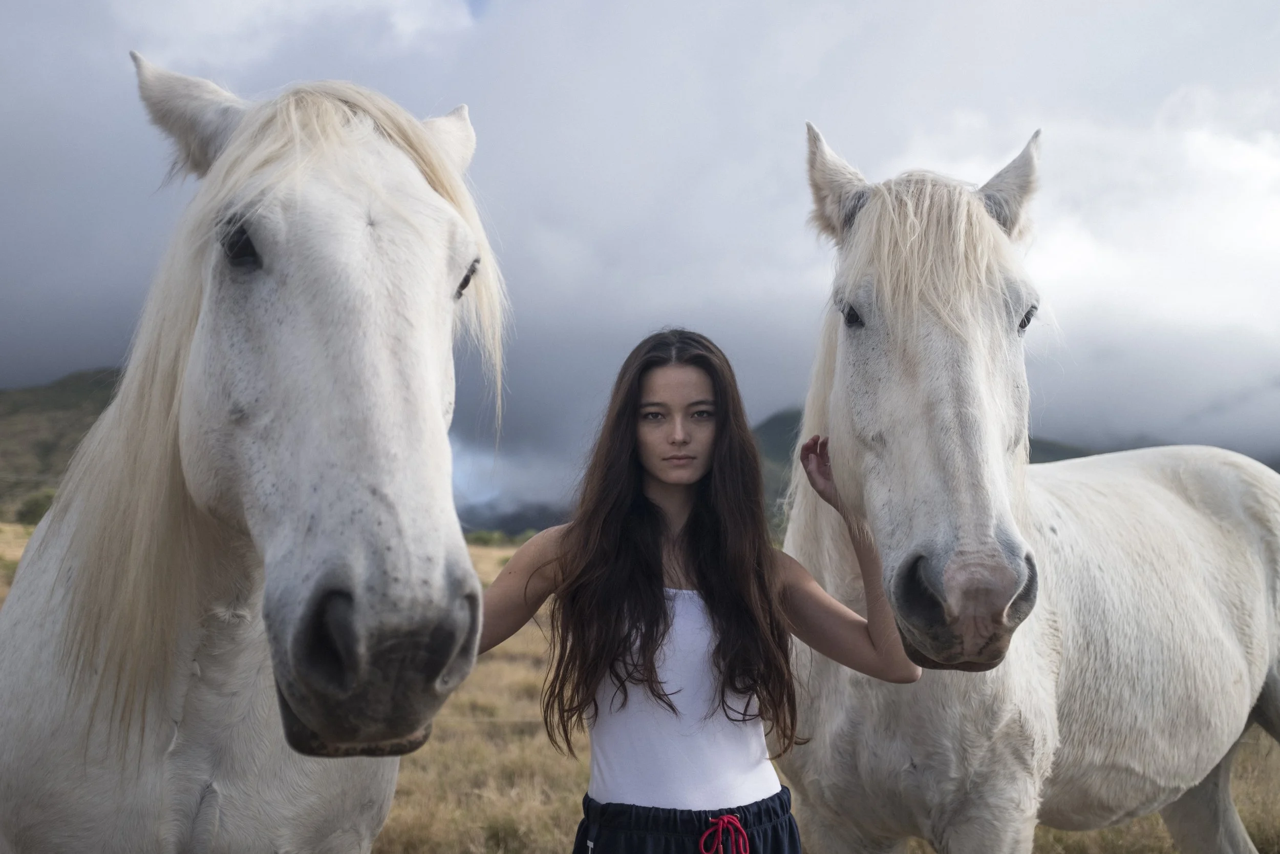 Une jeune femme avec de longs cheveux bruns, portant un débardeur blanc et un pantalon à la taille rouge, se tient entre deux chevaux blancs dans un paysage rural avec des montagnes et un ciel nuageux en arrière-plan.