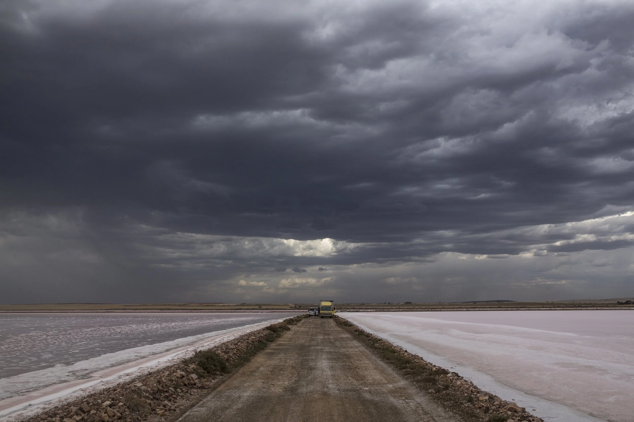 Un chemin de terre longeant un lac salé ou une étendue d'eau salée avec des montagnes de sel, sous un ciel nuageux sombre.