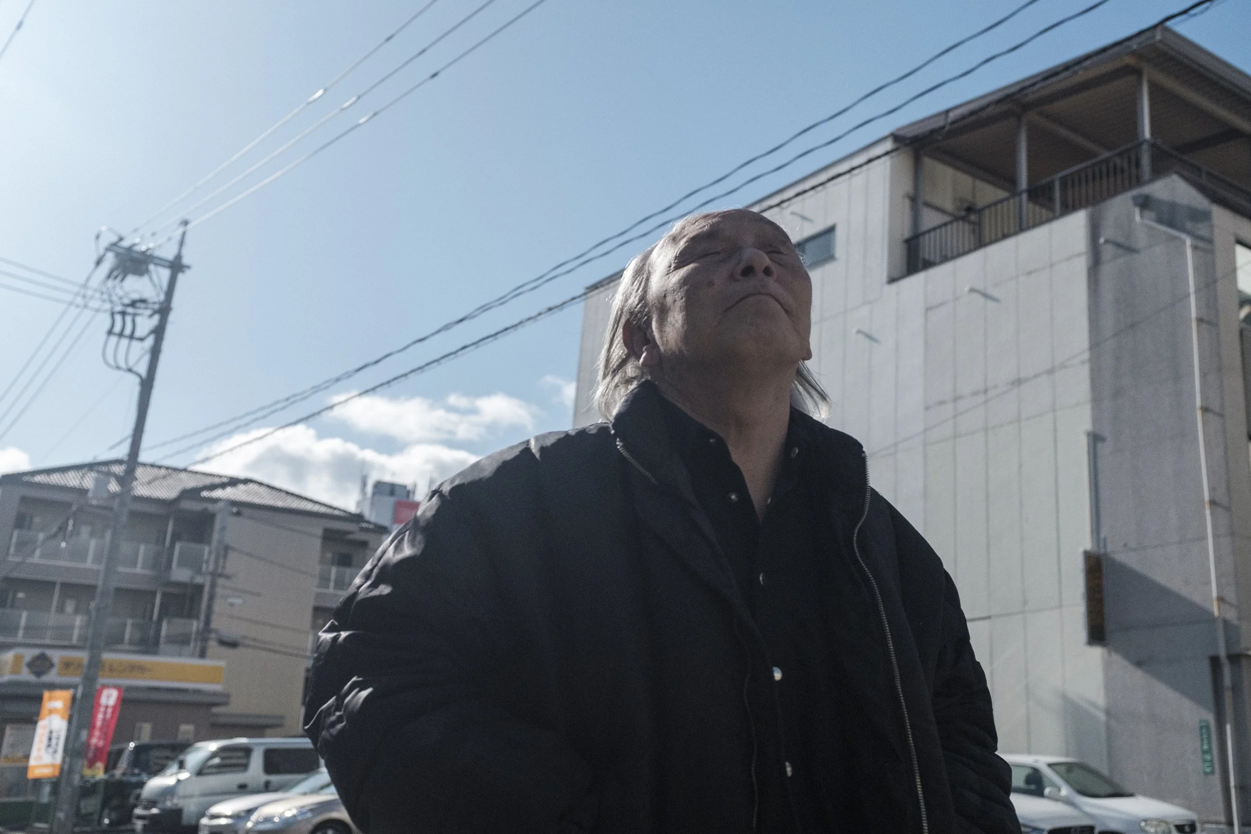 Homme âgé portant une veste noire, regardant vers le haut sous un ciel ensoleillé.