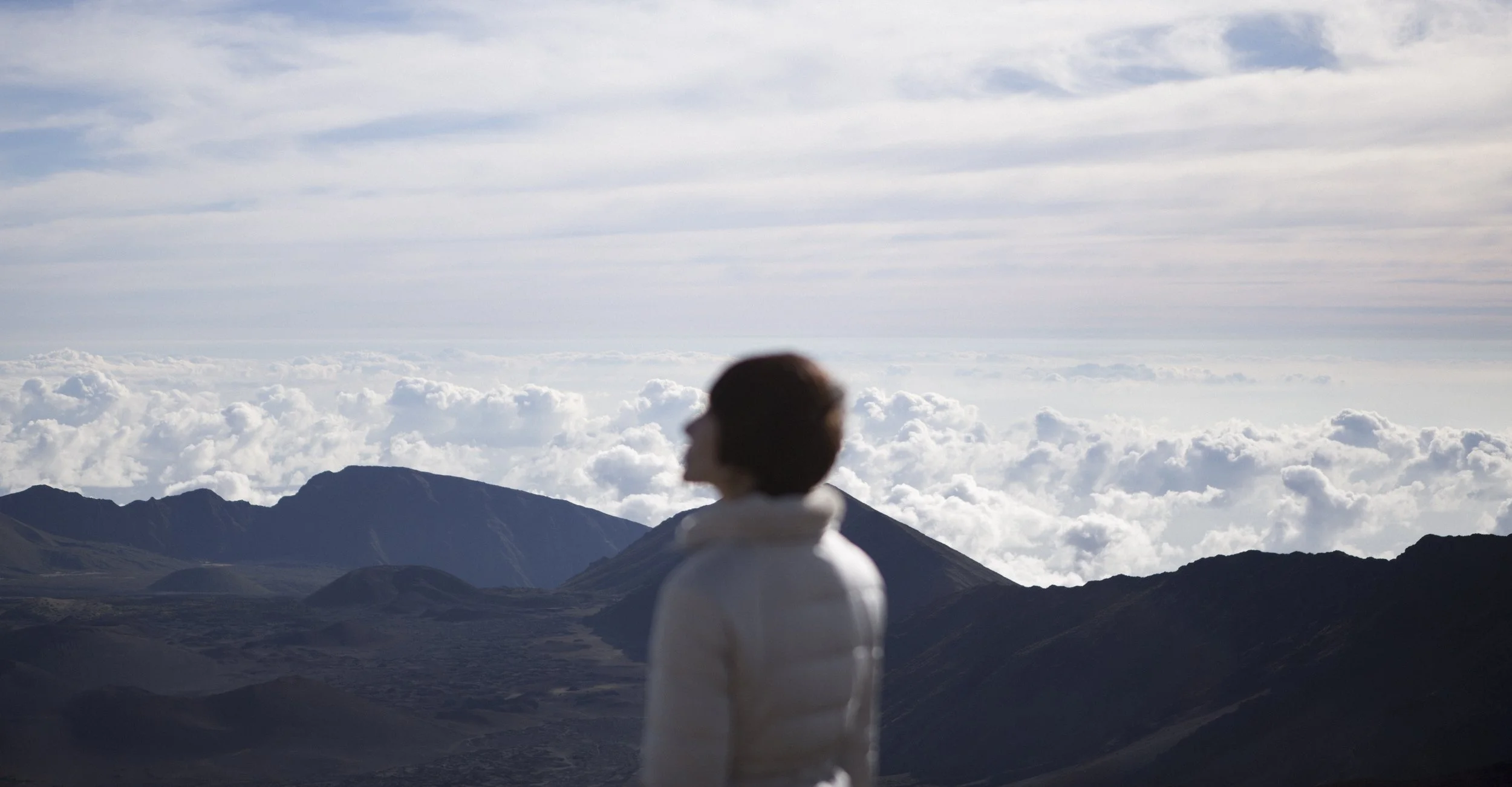 Une personne regarde un paysage de montagnes et de nuages en arrière-plan, vêtue d'une veste blanche, dans un environnement naturel magnifique.