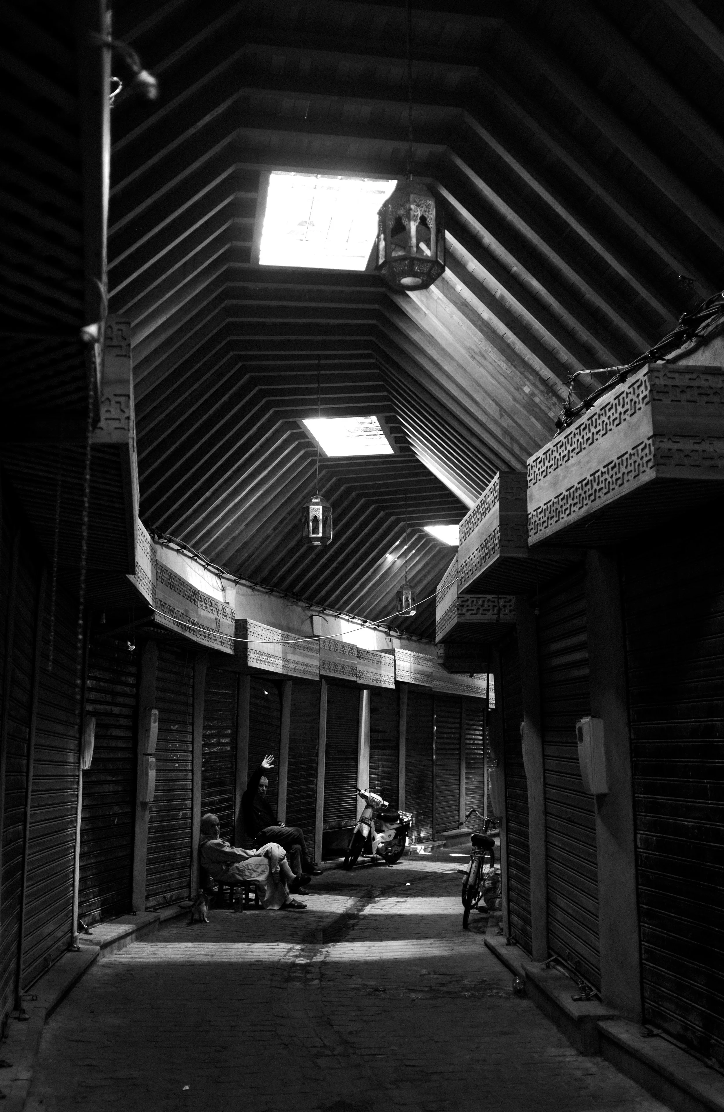 Rue intérieure avec trois personnes assises, entourée de boutiques fermées à roulettes, lumières suspendues et deux vélos, sous un plafond en bois avec fenêtres de toit.
