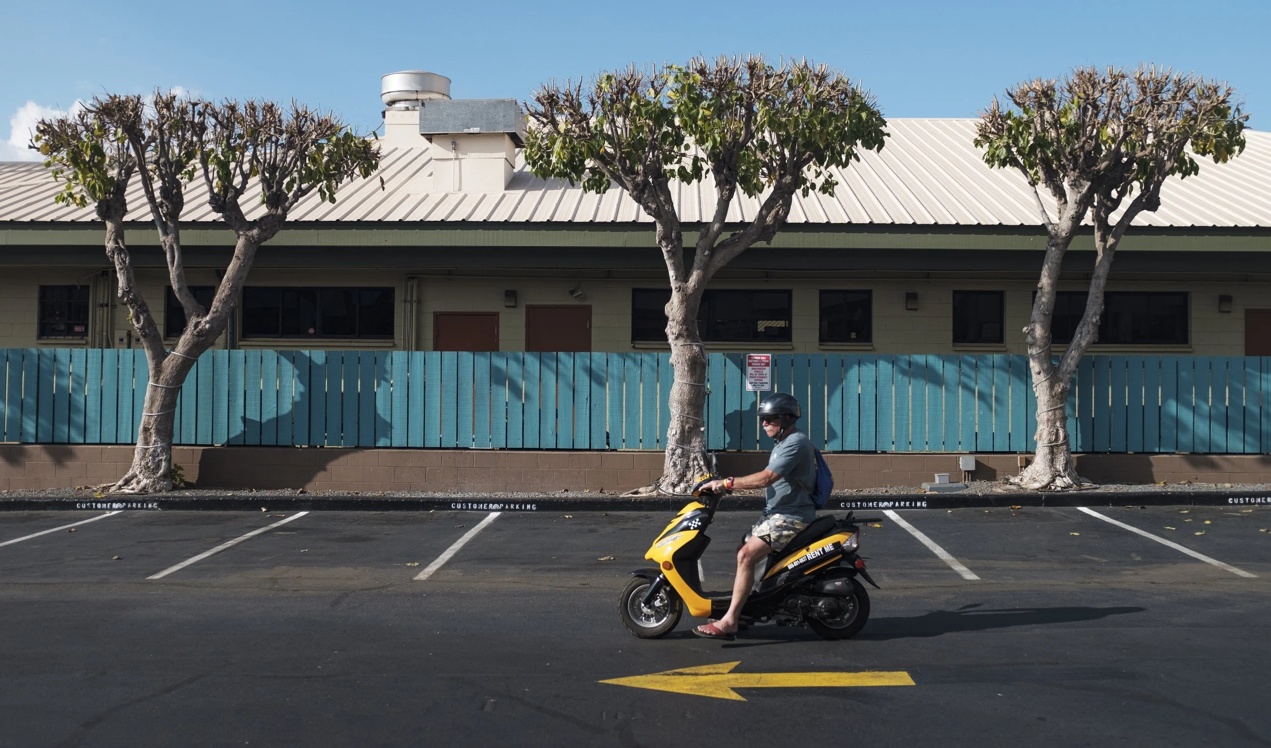 Un homme portant un casque, en t-shirt gris, shorts et sandales, roule sur un scooter jaune dans un parking vide, avec trois arbres taillés en arrière-plan et un bâtiment avec un mur bleu en arrière-plan.
