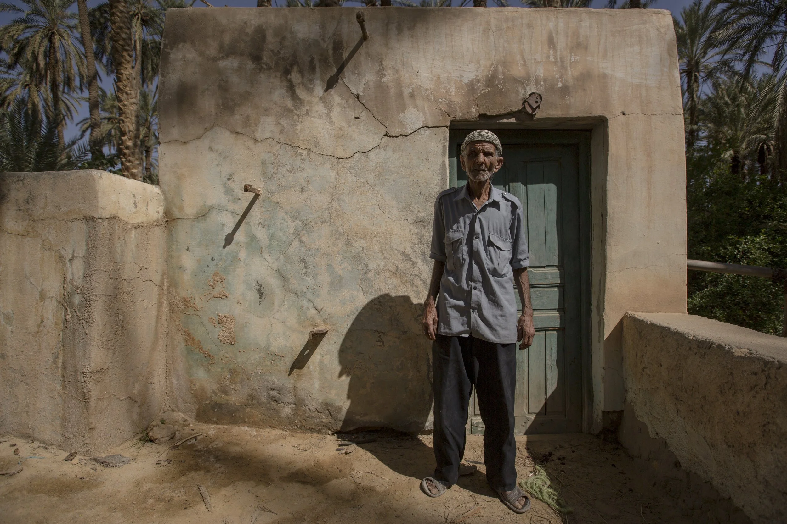 Un homme âgé se tient devant une porte d'une maison en adobe, avec des palmiers en arrière-plan, sous un ciel clair et ensoleillé.