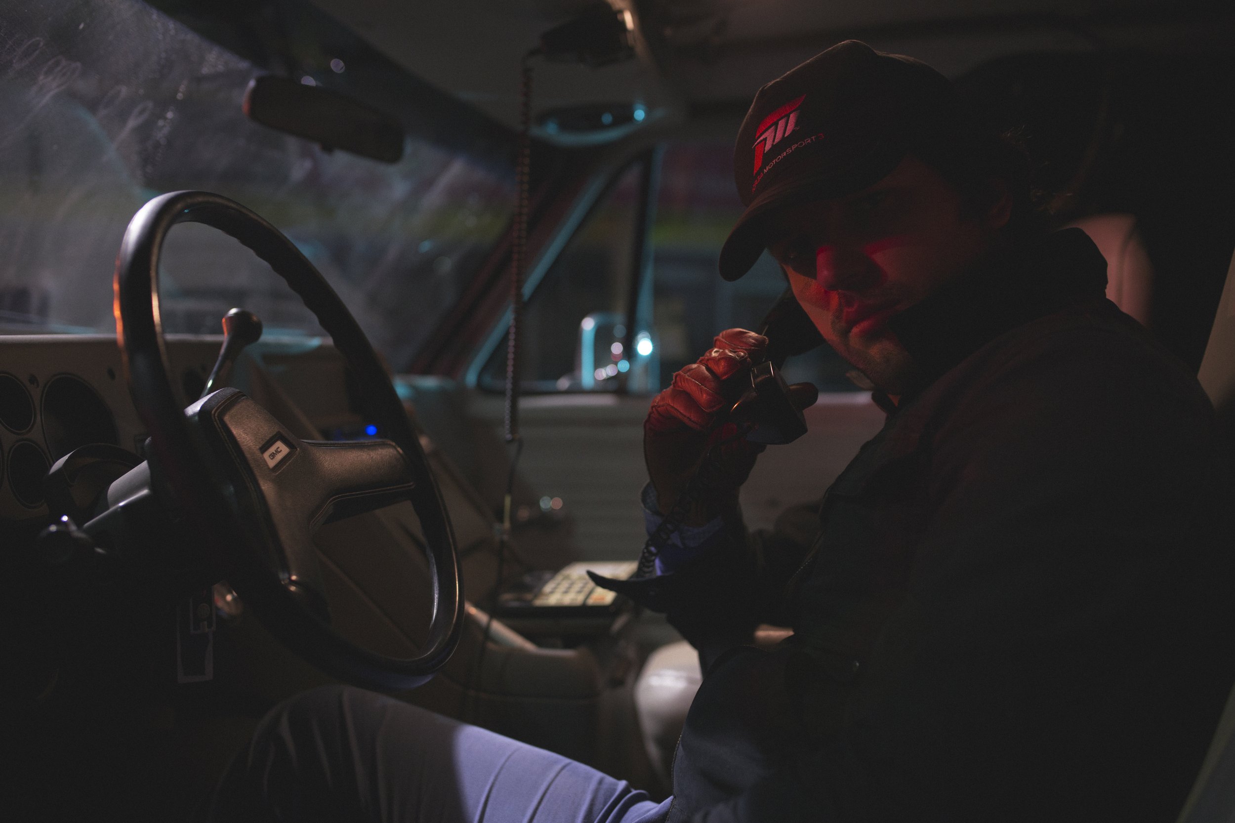 Un homme dans l'habitacle d'une voiture, portant une casquette noire et des gants, est en train de parler dans un talkie-walkie. Il semble être dans une situation de nuit ou dans un environnement sombre.