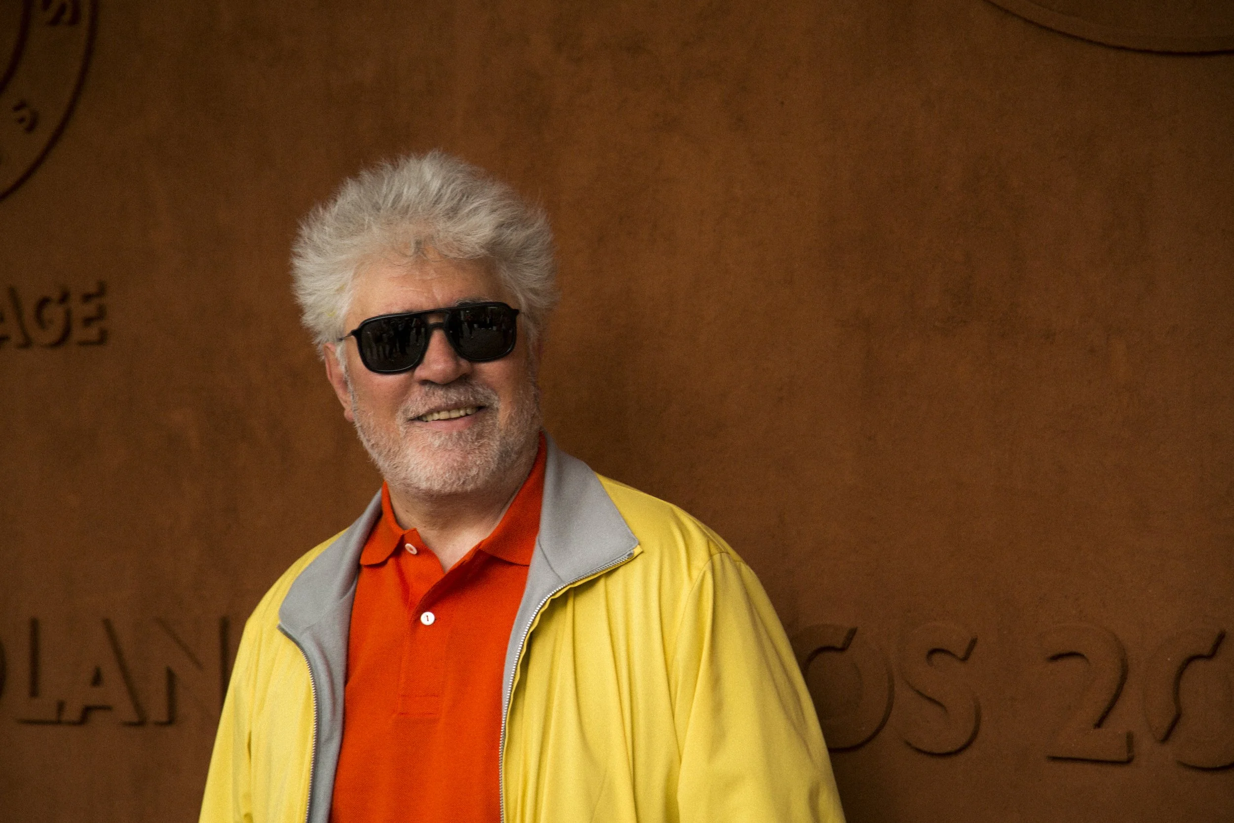Un homme avec des cheveux gris, portant des lunettes de soleil noires, un chemisier orange, une veste grise avec un col orange, et une veste jaune, souriant devant un fond marron avec des écritures et des dessins.