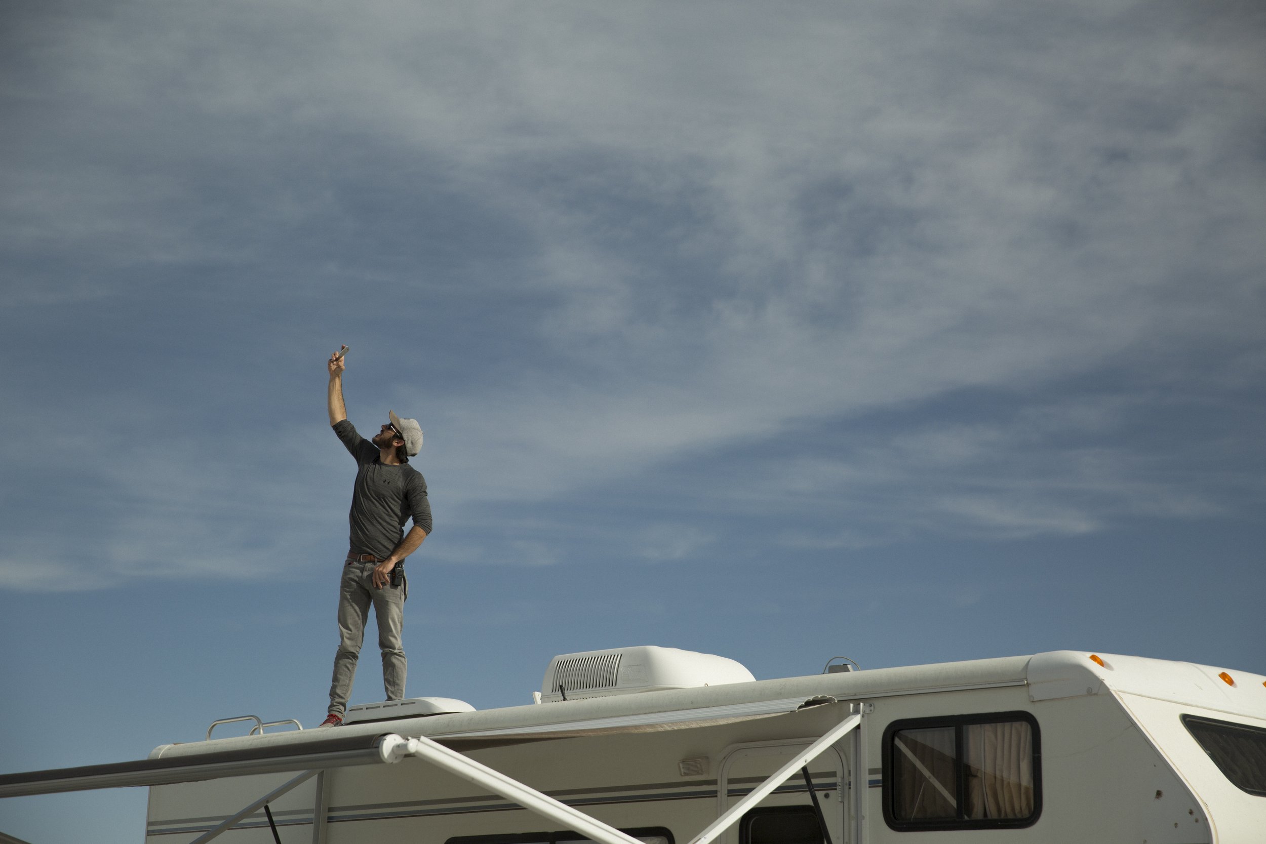 Un homme debout sur le toit d'une caravane, tenant un téléphone portable en l'air, regardant vers le haut, avec un ciel nuageux en arrière-plan.