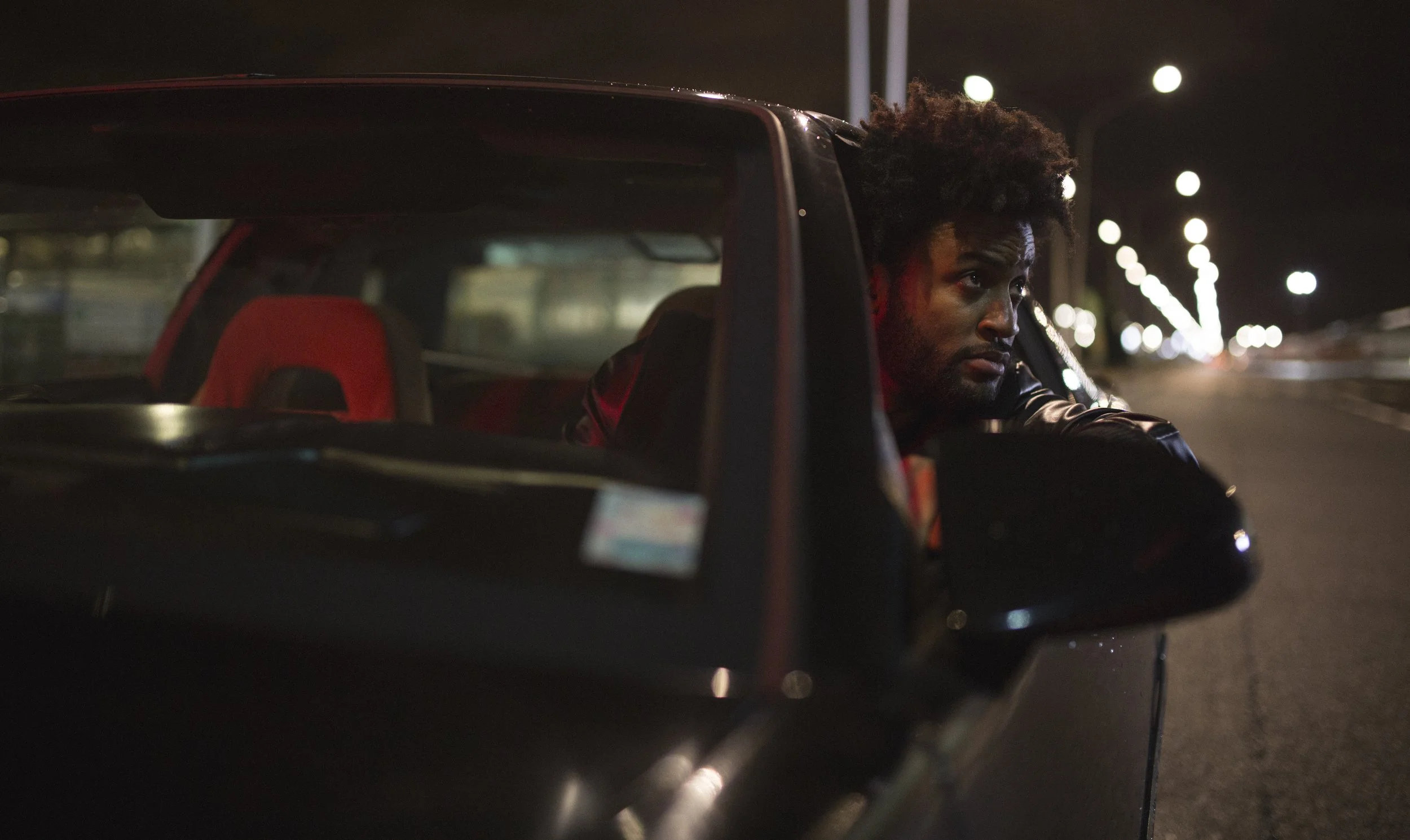 Un homme semble pensif, assis dans une voiture de nuit, avec des lumières de rue floues en arrière-plan lors d'une sortie nocturne.
