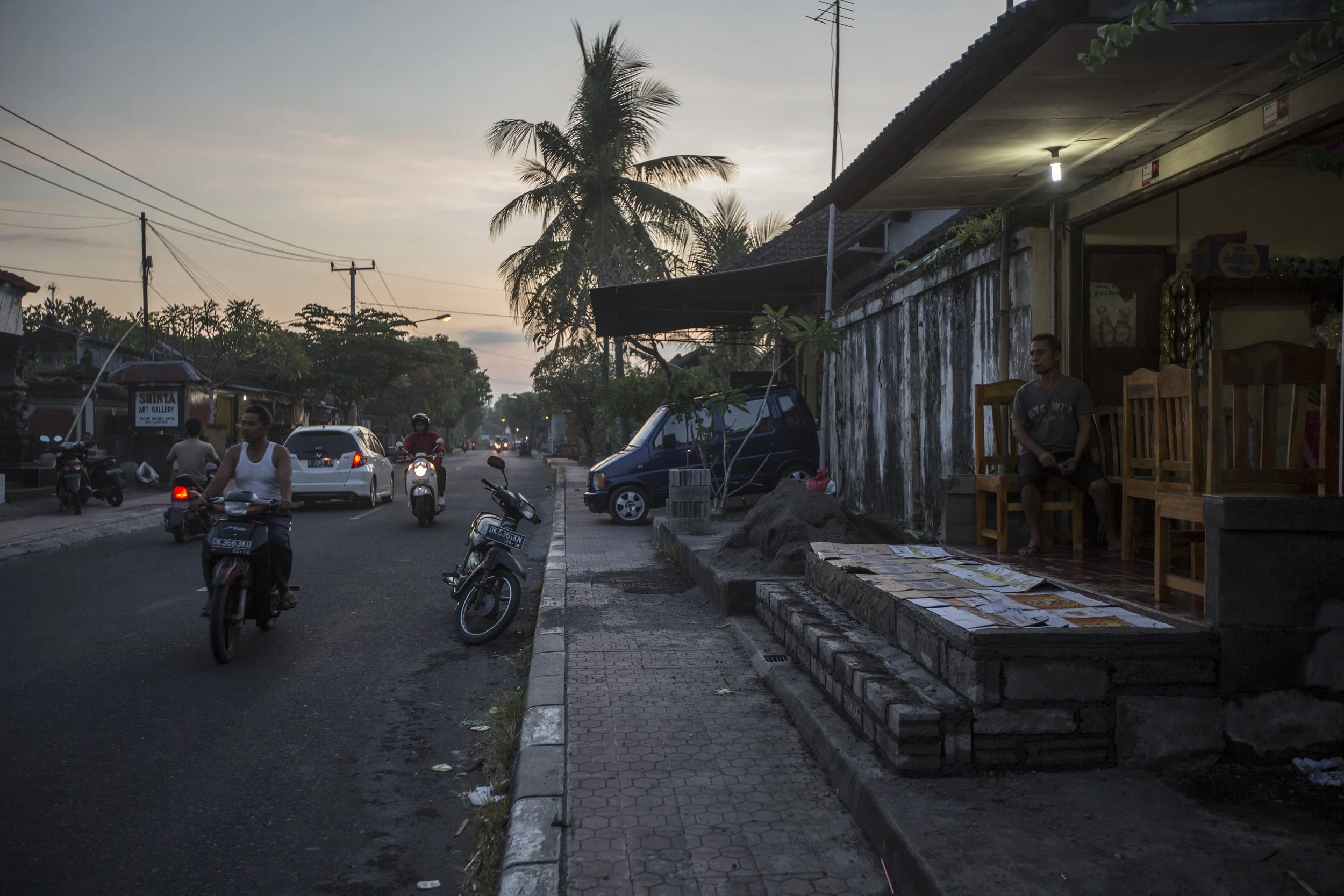 Rue urbaine en fin d'après-midi avec des motos et voitures en mouvement, un homme assis devant une boutique, palmiers et ciel nuageux en arrière-plan.