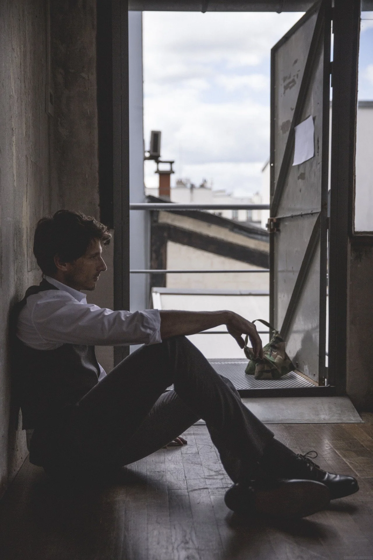Un homme assis par terre, appuyé contre un mur intérieur, à côté d'une porte ouverte donnant sur un balcon avec un ciel nuageux en arrière-plan.