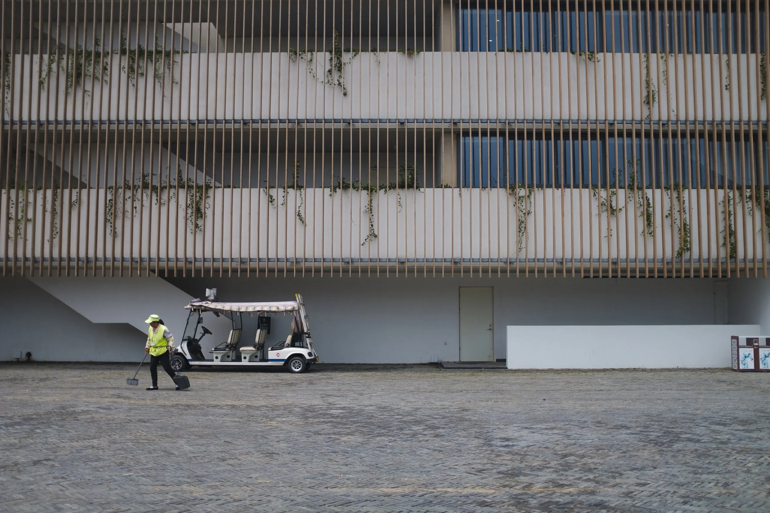Une personne en vestiaire jaune et casque vert, utilisant un balai pour nettoyer la zone pavée. Un véhicule de golf blanc avec un toit multicolore stationne à proximité d'un bâtiment moderne avec des balustrades en bois et un mur blanc avec des plant