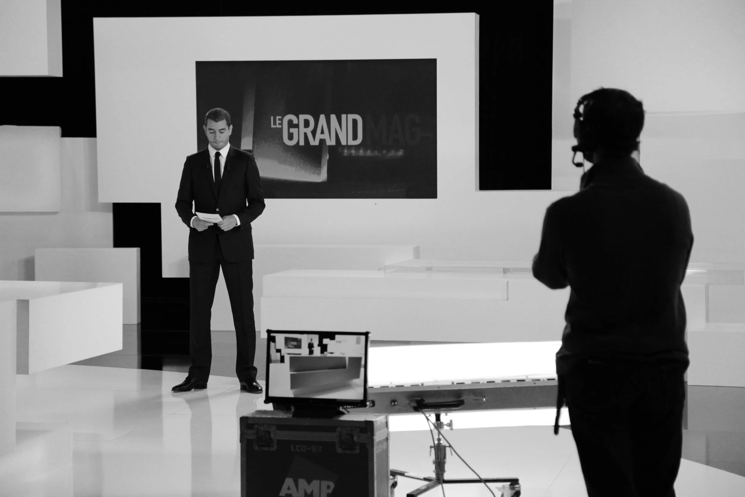 Une scène de studio de télévision en noir et blanc avec un homme en costume tenant des papiers, et une personne avec un casque et un micro regardant vers lui, derrière un équipement de caméra.