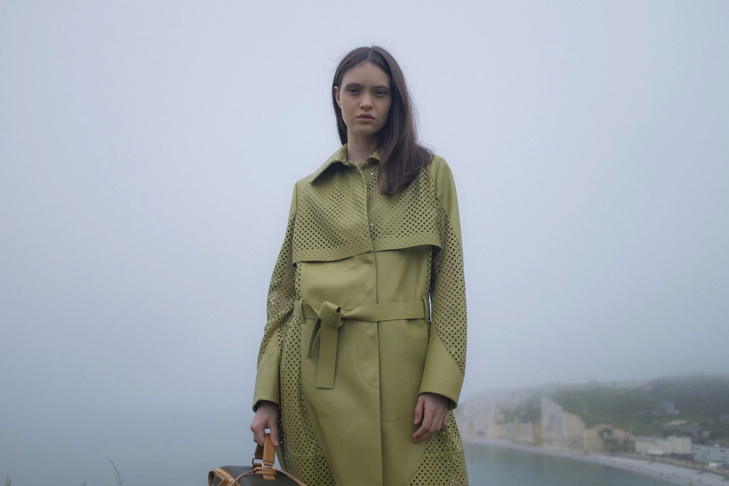 Une jeune femme avec des cheveux bruns, portant un trench-coat vert olive avec des motifs en losange, se tenant devant un paysage côtier brumeux.