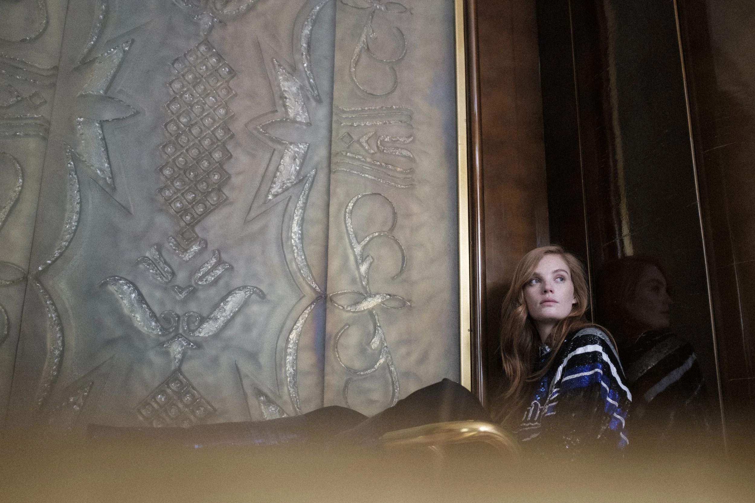 Jeune femme aux cheveux roux assise dans un espace intérieur, recouverte d'un vêtement à rayures noires, blanches et bleues, regardant vers la gauche. Elle est assise près d'un mur en bois foncé avec une surface réfléchissante. Une grande décoration 