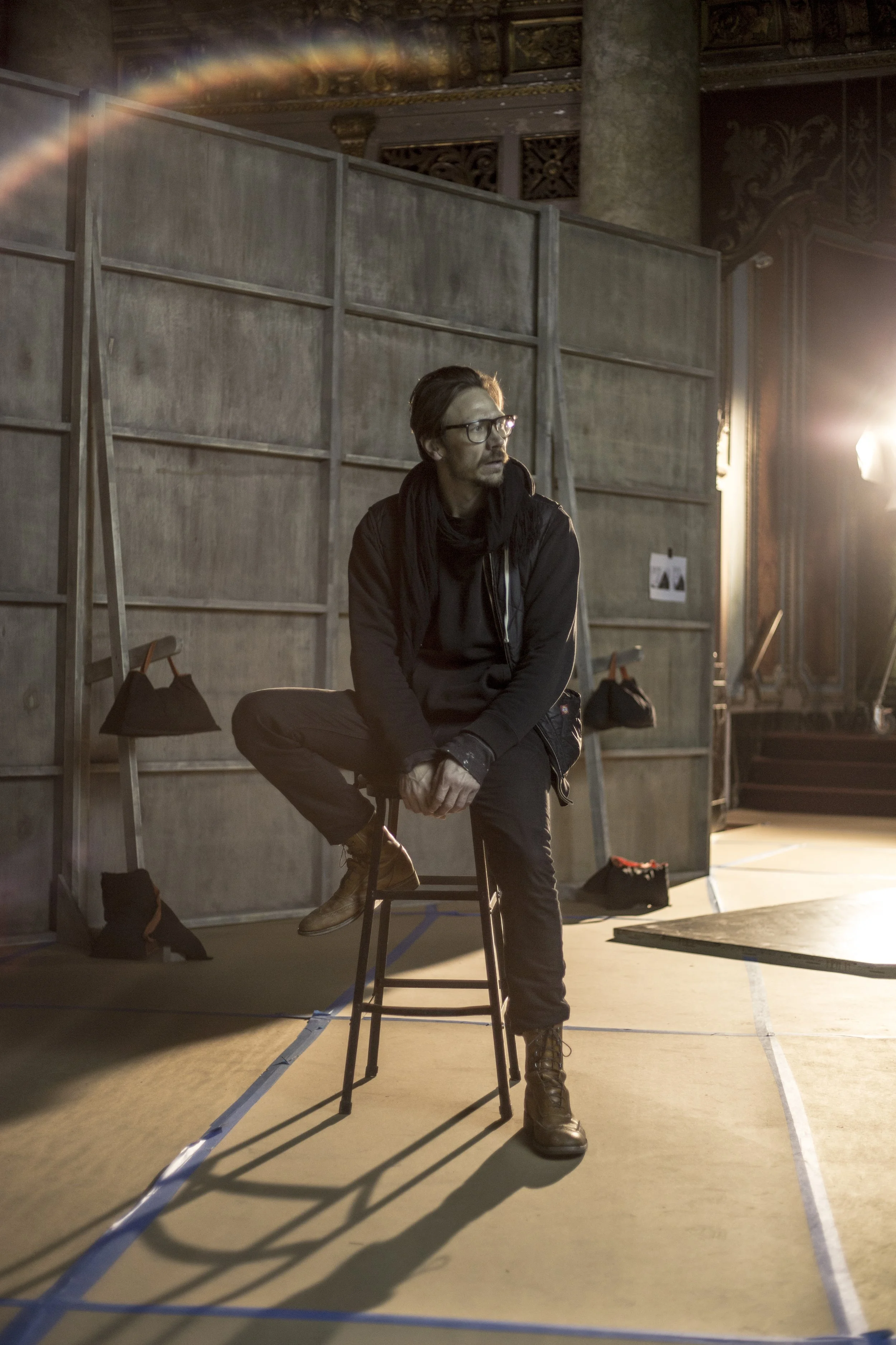 Un homme assis sur une chaise dans un studio, portant des lunettes et un vêtement noir, avec un éclairage de studio brillant à droite et un décor en arrière-plan.