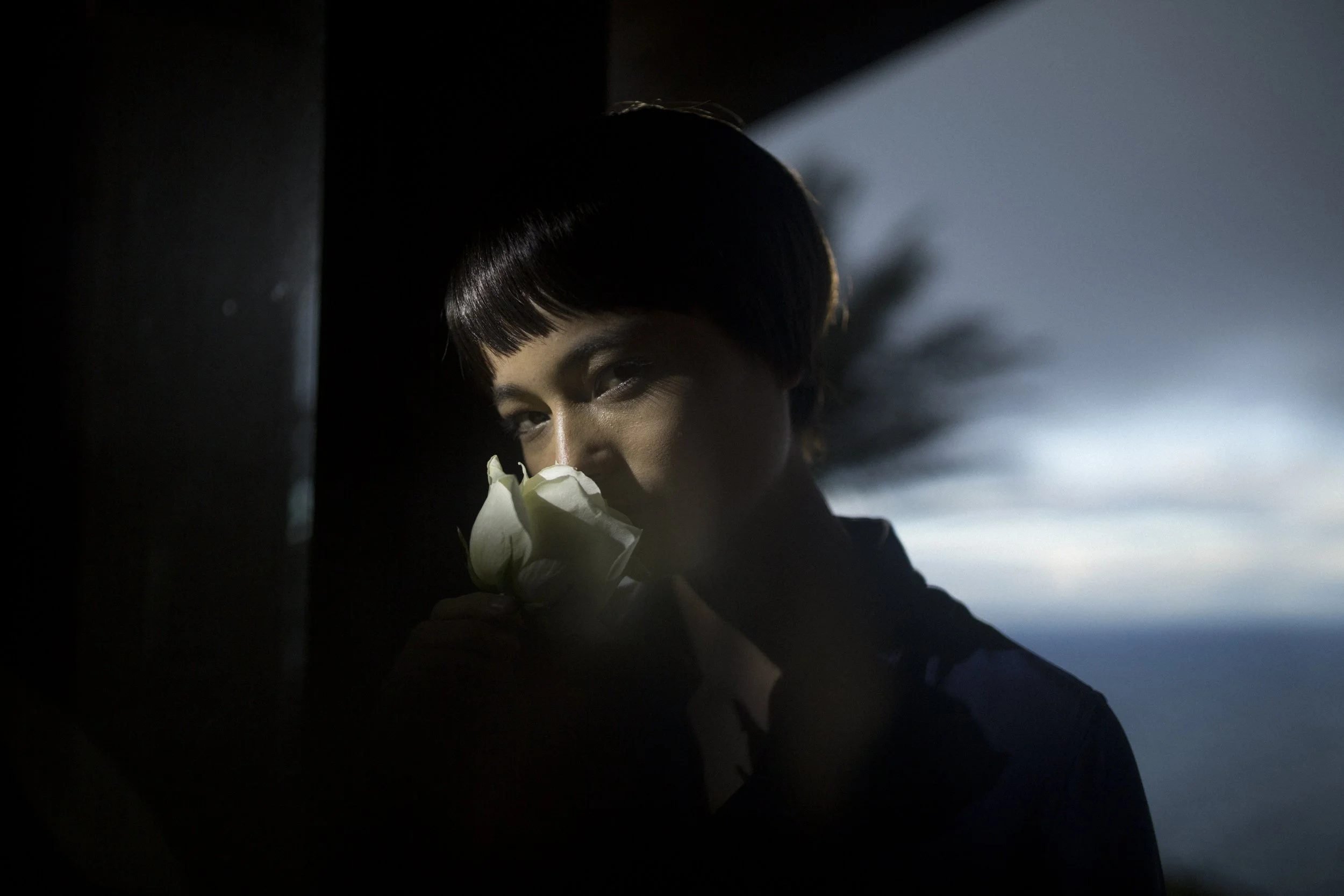 Femme avec une coupe au carré noir, tenant un bouquet de fleurs blanches, dans un environnement sombre avec un arrière-plan de ciel nuageux.