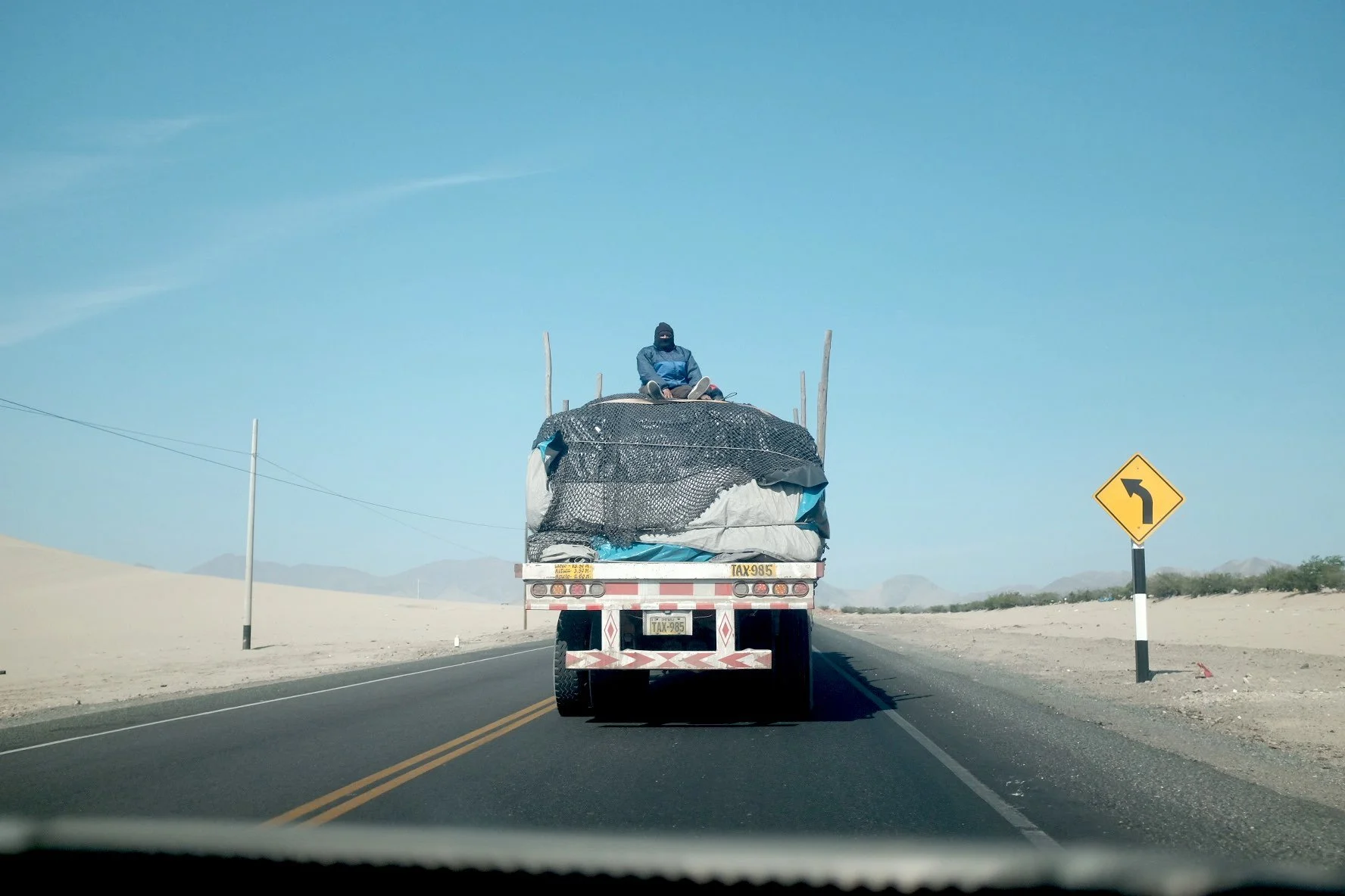 Un camion transportant des sacs et du matériel, vu de derrière, conduisant sur une route en zone désertique avec un panneau de virage à gauche.