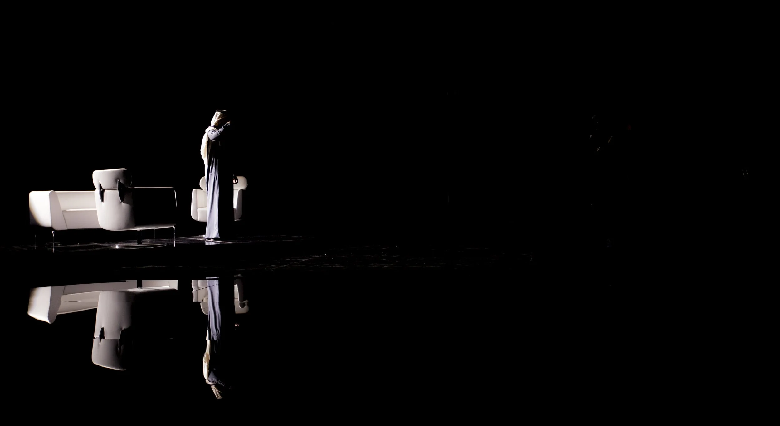 Une personne habillée de façon traditionnelle, portant un hijab, debout à côté de canapés blancs, dans un environnement très sombre avec un reflet au sol.