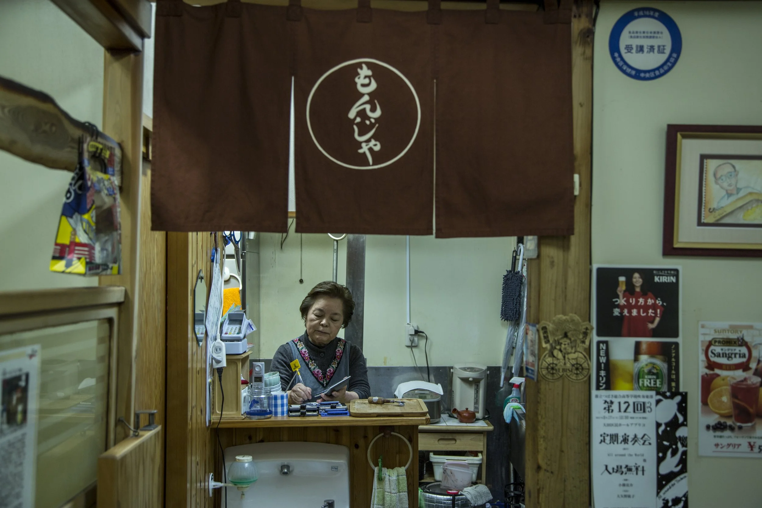 Une femme âgée derrière un comptoir dans un petit restaurant japonais, tenant un carnet ou un menu, avec diverses bouteilles, appareils et objets de cuisine autour d'elle, et un rideau marron avec des caractères japonais au-dessus.