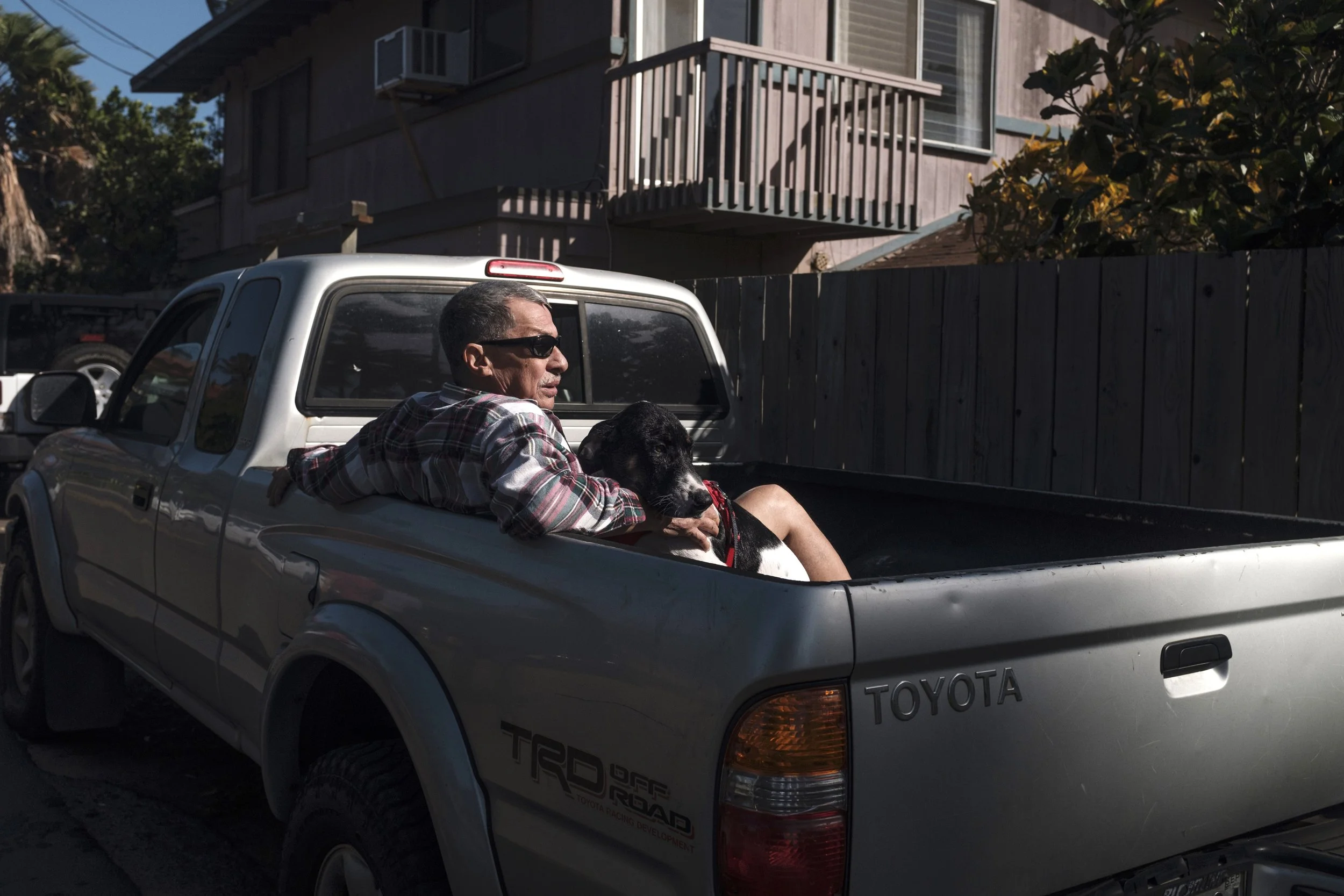 Un homme portant des lunettes de soleil et une chemise en flanelle repose dans la benne d'un camion Toyota avec un chien noir et blanc très proche, dans une zone résidentielle en plein jour.