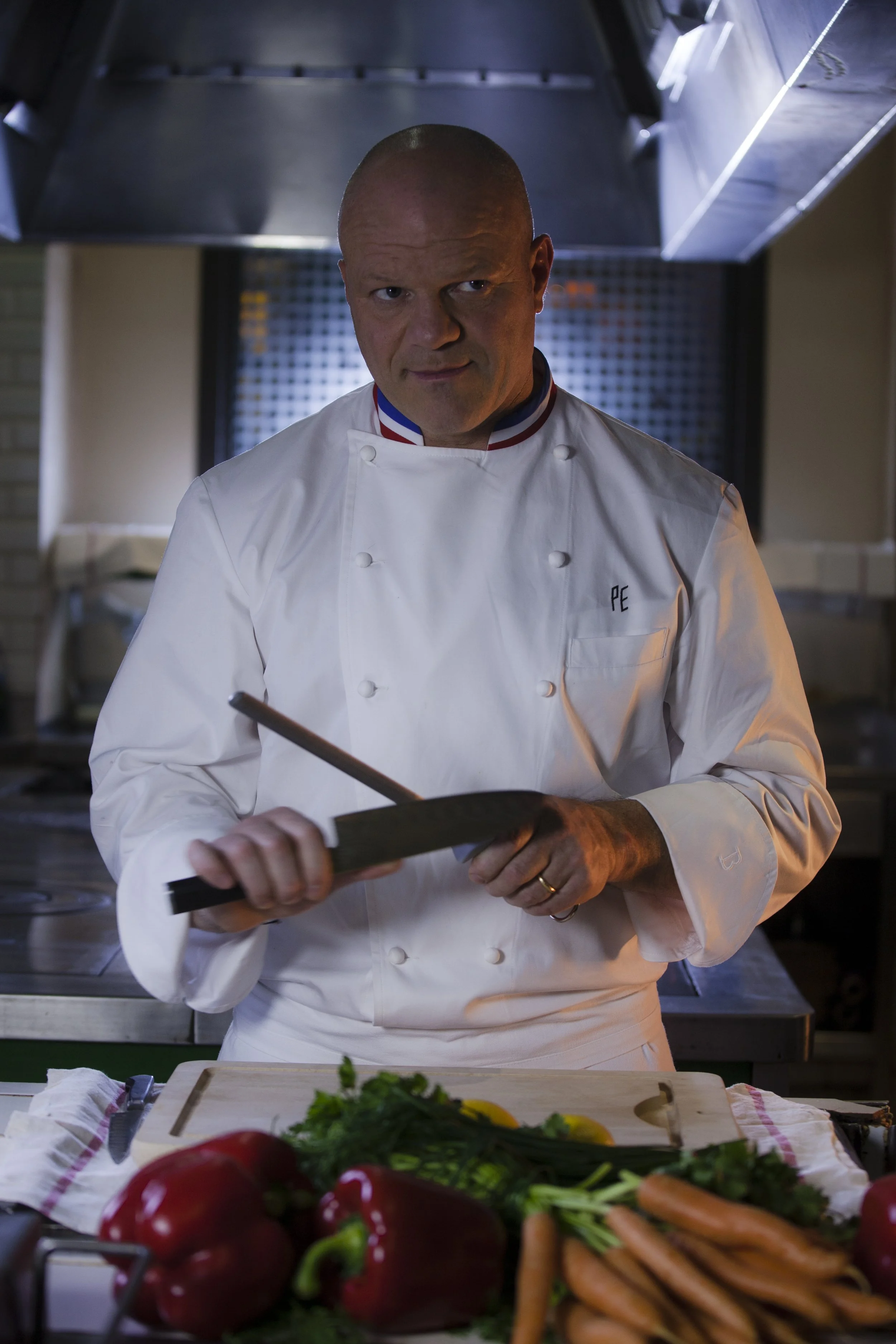 Chef professionnel en cuisine, portant une toque blanche, tenant un couteau, avec des légumes frais sur la table en face.